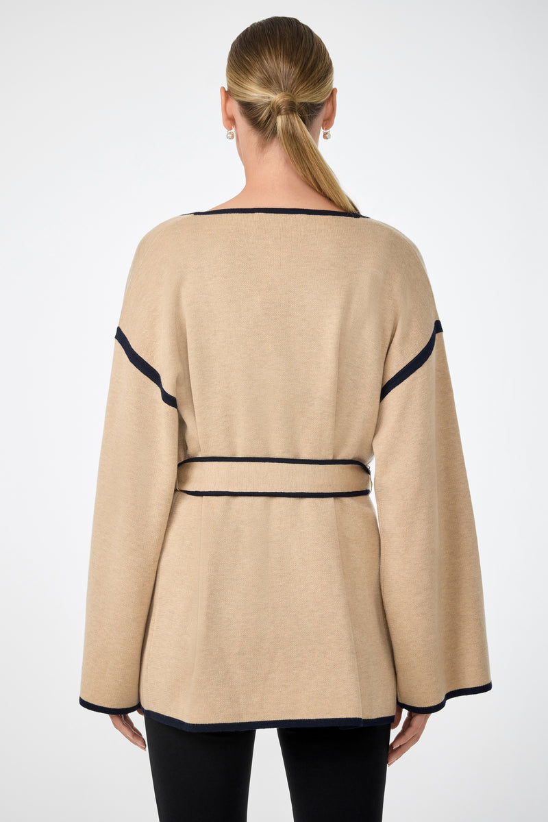 A person with straight, light brown hair in a low ponytail is shown from behind, wearing the beige Wright Wrap Cardigan with black trim and belt, paired with black pants.