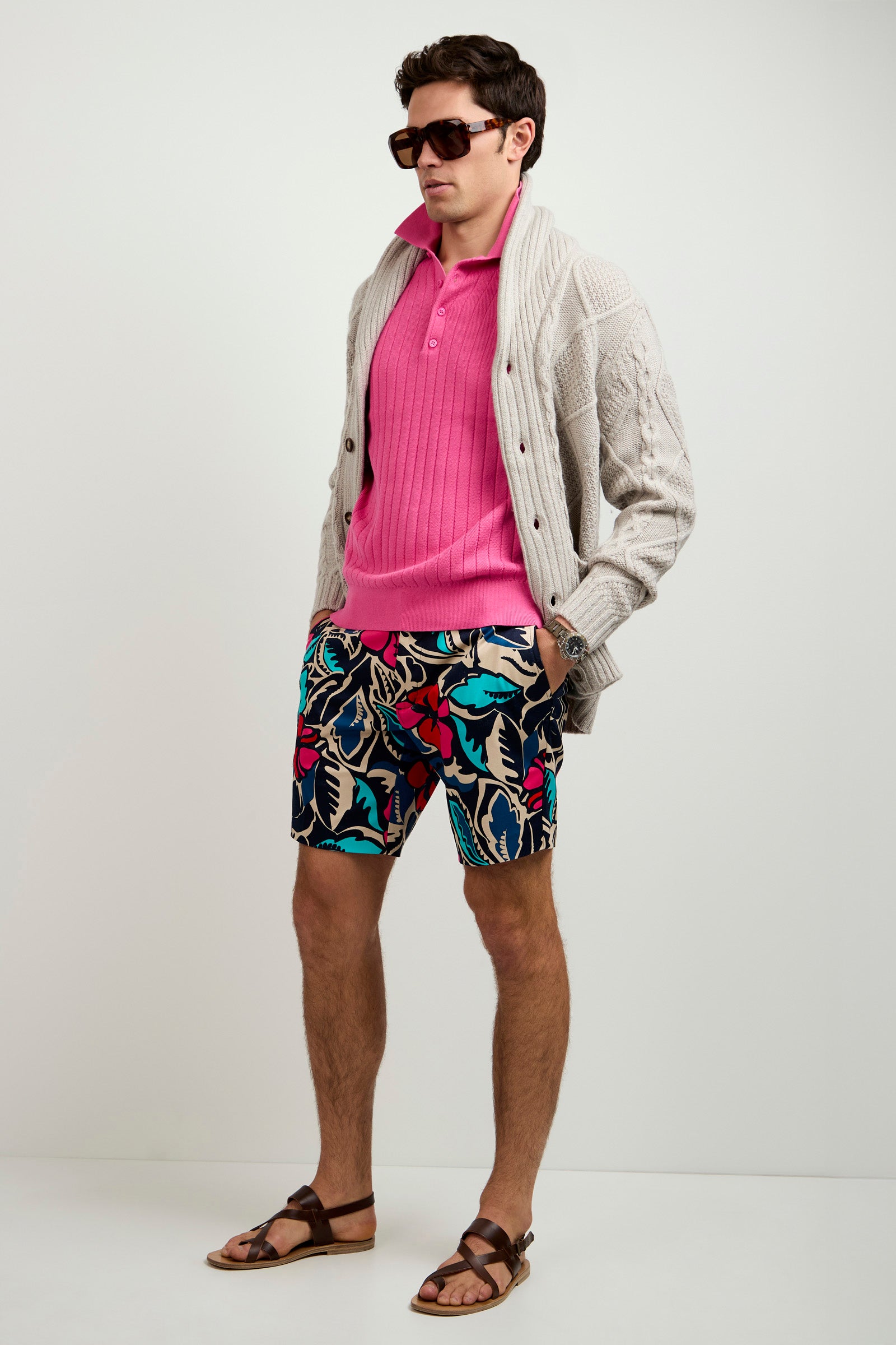 A man wearing sunglasses, a pink polo shirt, colorful floral shorts, brown sandals, and the Brighton Cardigan stands against a plain background.