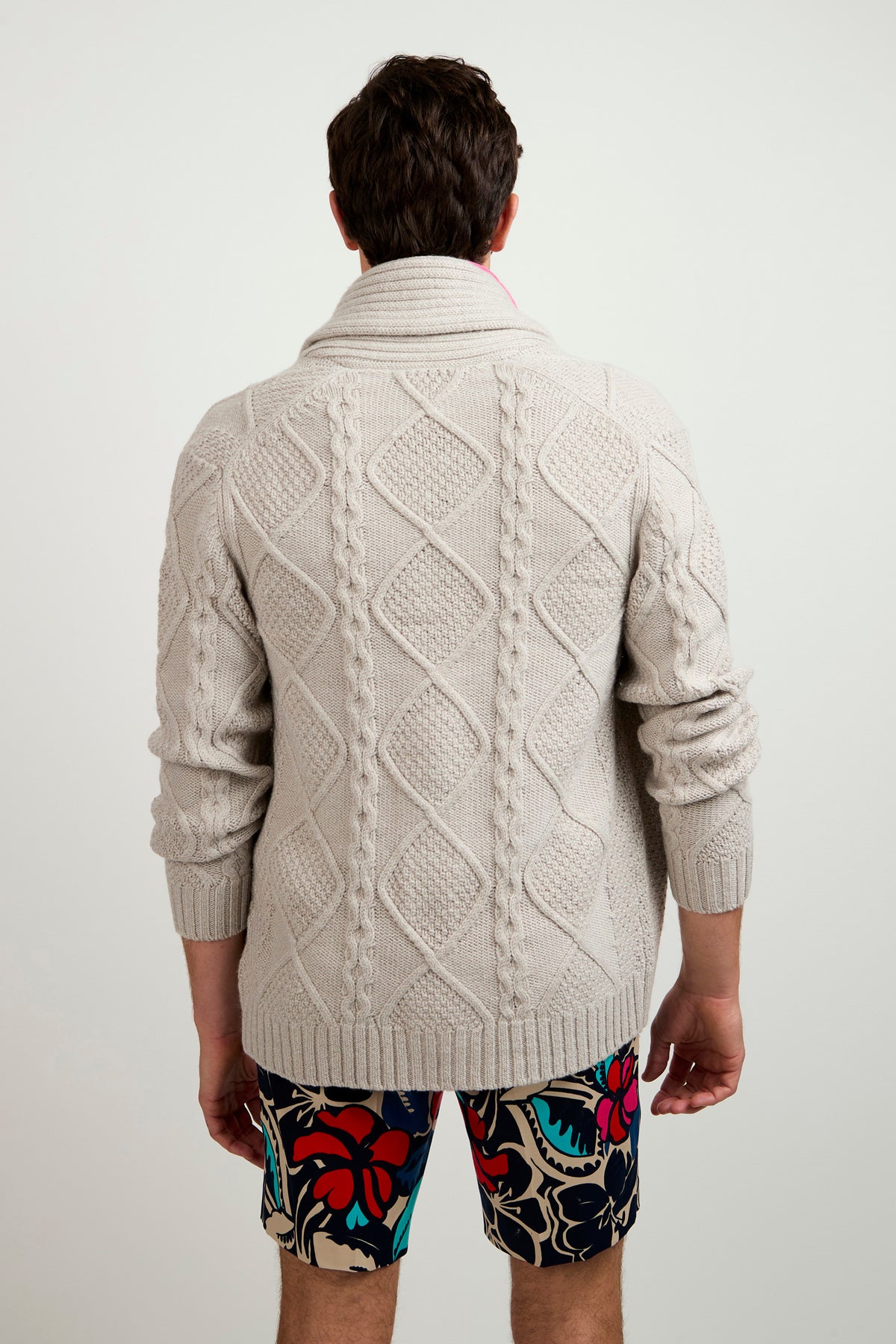 A man stands facing away, wearing the Brighton Cardigan—a light gray, patterned knit sweater—and colorful floral shorts against a plain white background.