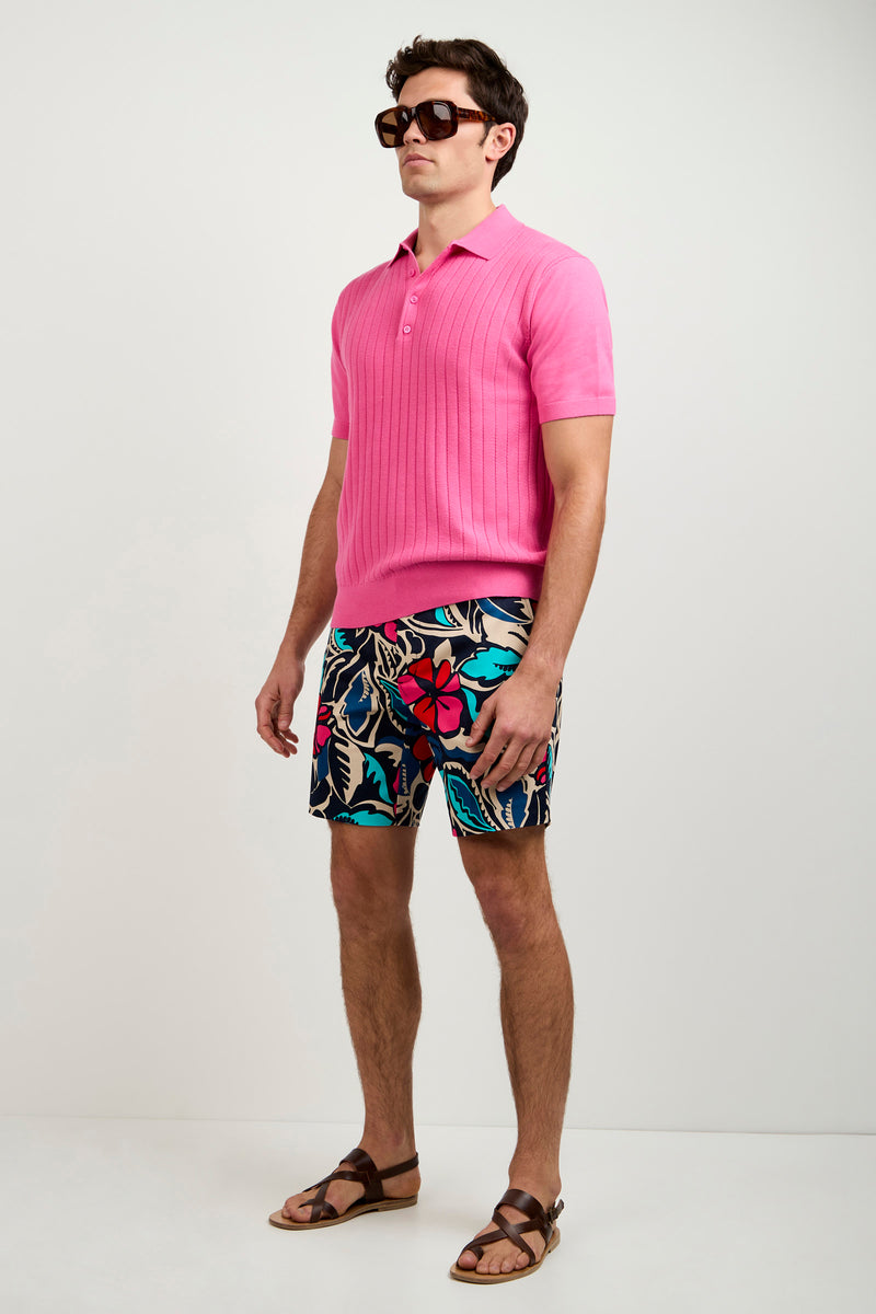 A man wearing the Alvarez Short-Sleeve Polo in bright pink, paired with colorful floral shorts, brown sandals, and large dark sunglasses stands against a plain light background.