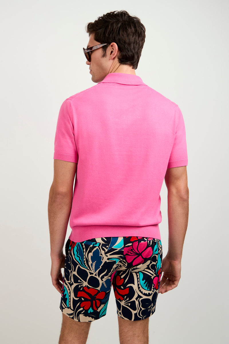 A man with short brown hair and sunglasses, wearing the bright pink Alvarez Short-Sleeve Polo and colorful floral shorts, stands facing away from the camera against a plain light background.