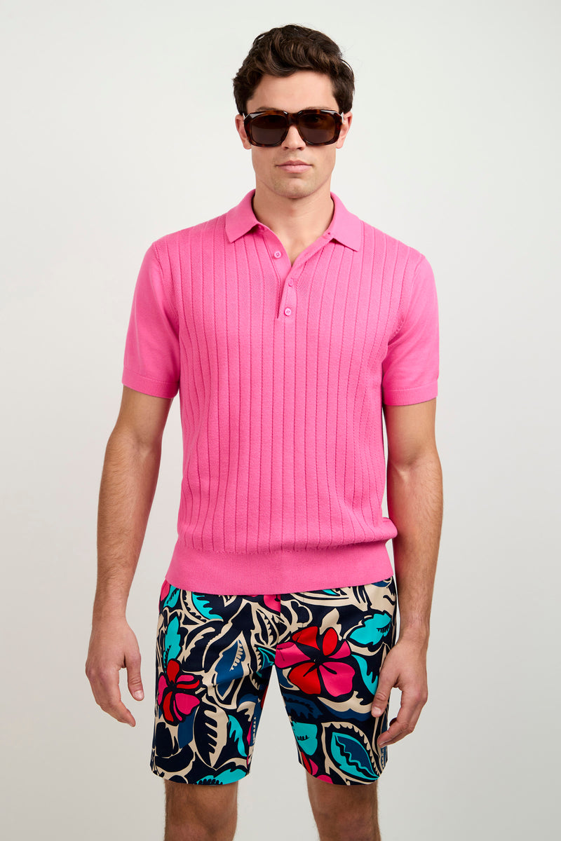 A person in an Alvarez Short-Sleeve Polo, floral patterned shorts, and oversized dark sunglasses stands against a plain light background.