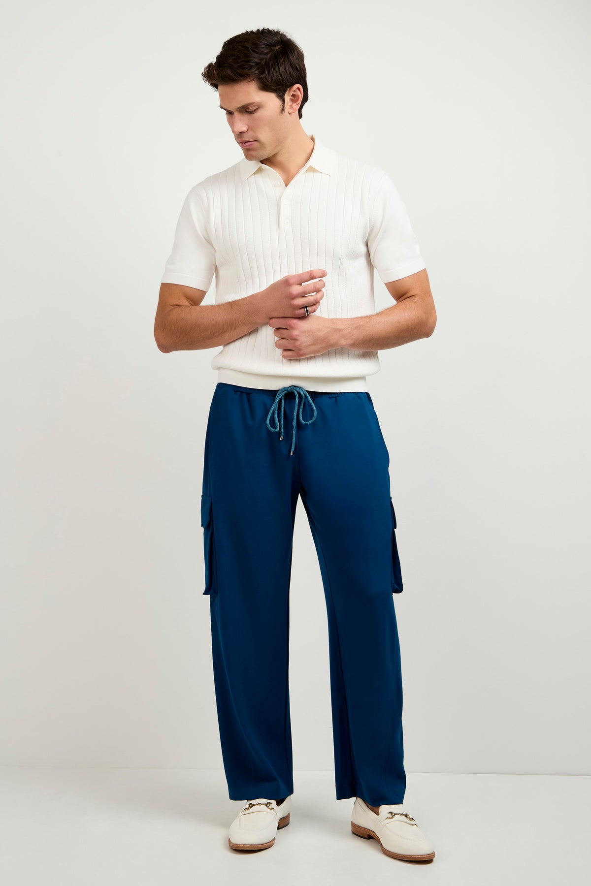 A man stands against a plain light background, wearing the Alvarez Short-Sleeve Polo in white, blue drawstring cargo pants, and white loafers. He looks down with his hands touching in front of him.