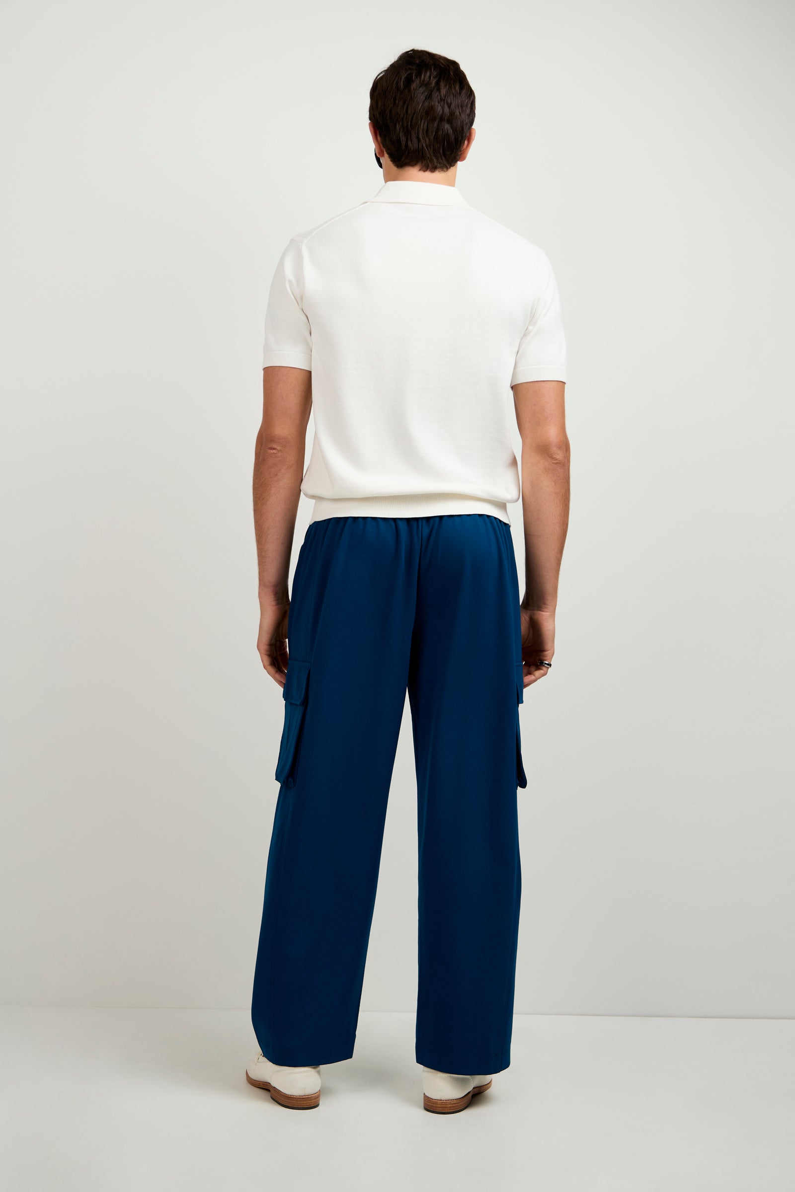 A man faces away from the camera, wearing a short-sleeve white shirt, white shoes, and Lounge Cargo Pant—loose blue pants with pockets—against a plain light background.