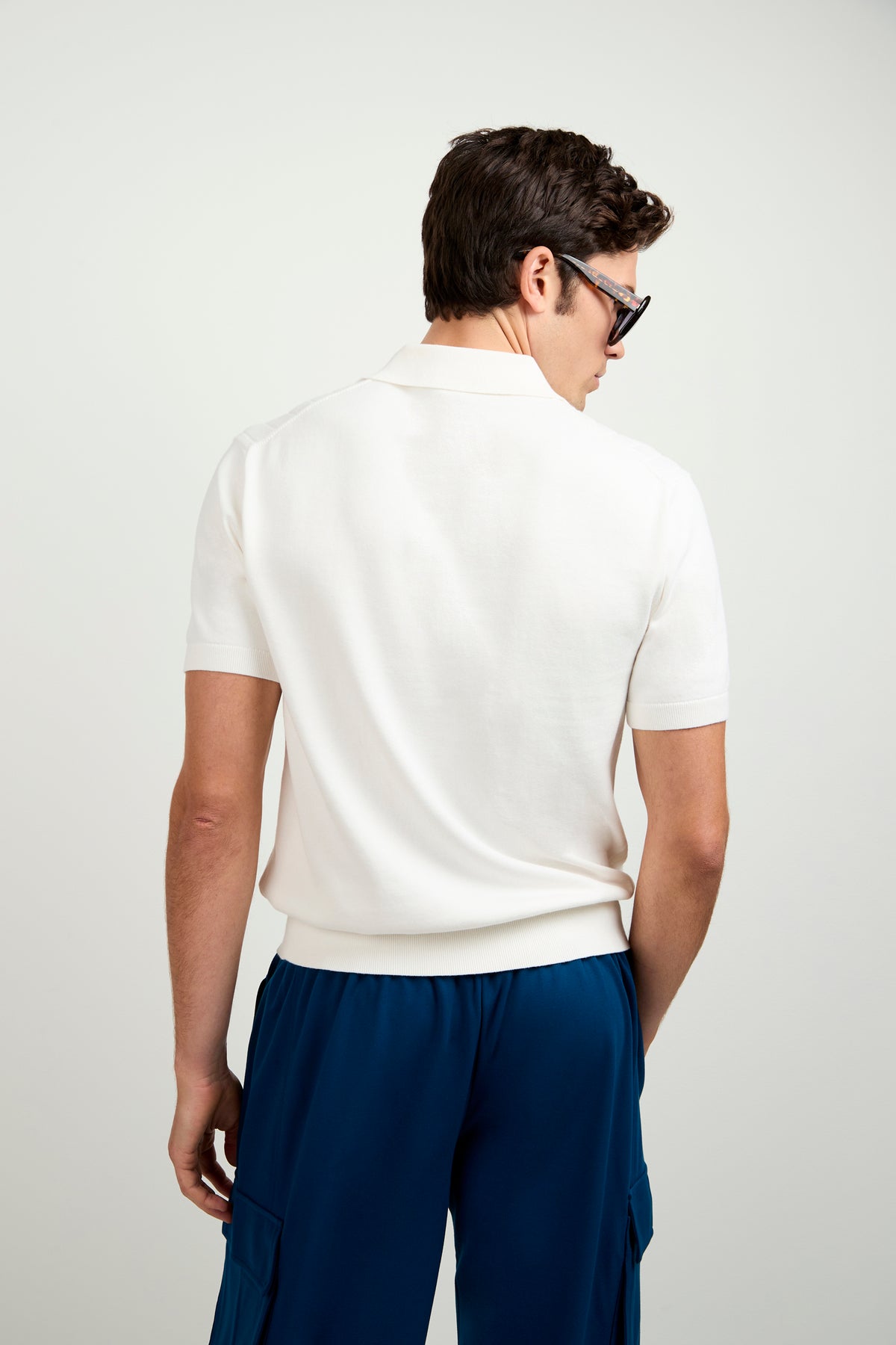 A man with short brown hair, in sunglasses and blue cargo pants, faces away from the camera wearing the Alvarez Short-Sleeve Polo against a plain light background.
