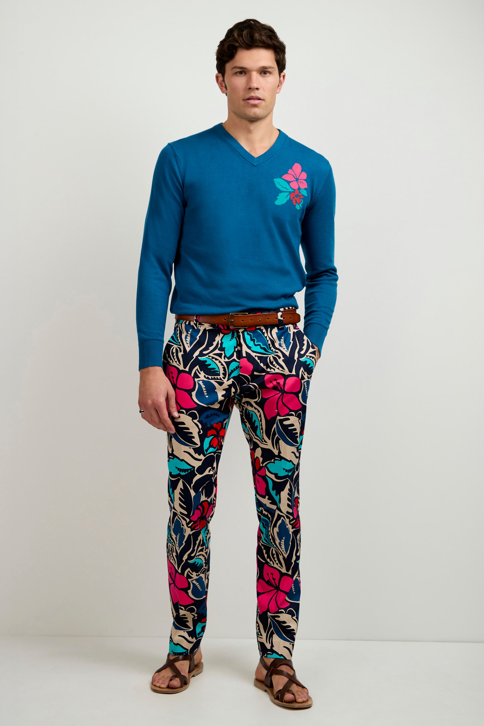 A man stands against a plain background wearing the Copacabana V-Neck Sweater, featuring a blue base with a pink flower design, paired with colorful floral print pants, a brown belt, and brown sandals. His hands are in his pockets as he faces the camera.
