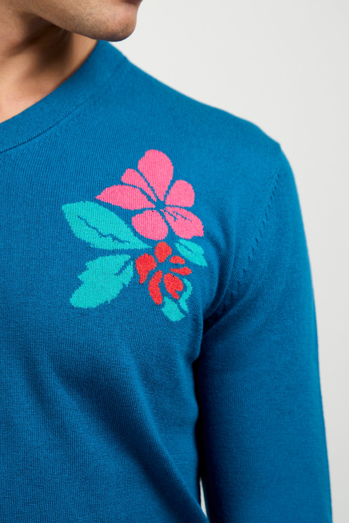 A person wearing the Copacabana V-Neck Sweater, featuring a blue color with pink and red floral design on the shoulder, is shown from neck to upper torso against a plain light background.