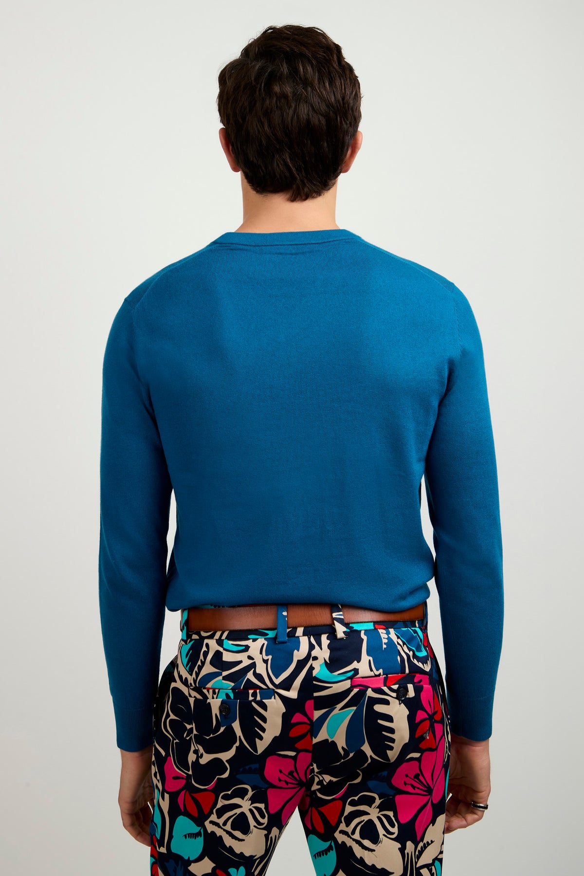 A person stands facing away, wearing the Copacabana V-Neck Sweater in bright blue and colorful, floral-patterned pants featuring red, black, cream, and teal on a light background. The backdrop is plain and light-colored.