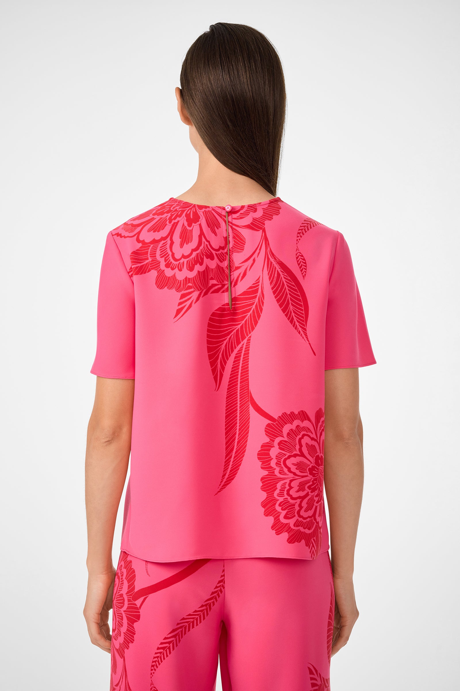 A woman with straight brown hair stands facing away, wearing the Sugarhill Top—a bright pink short-sleeve blouse with bold red floral patterns and matching pants.