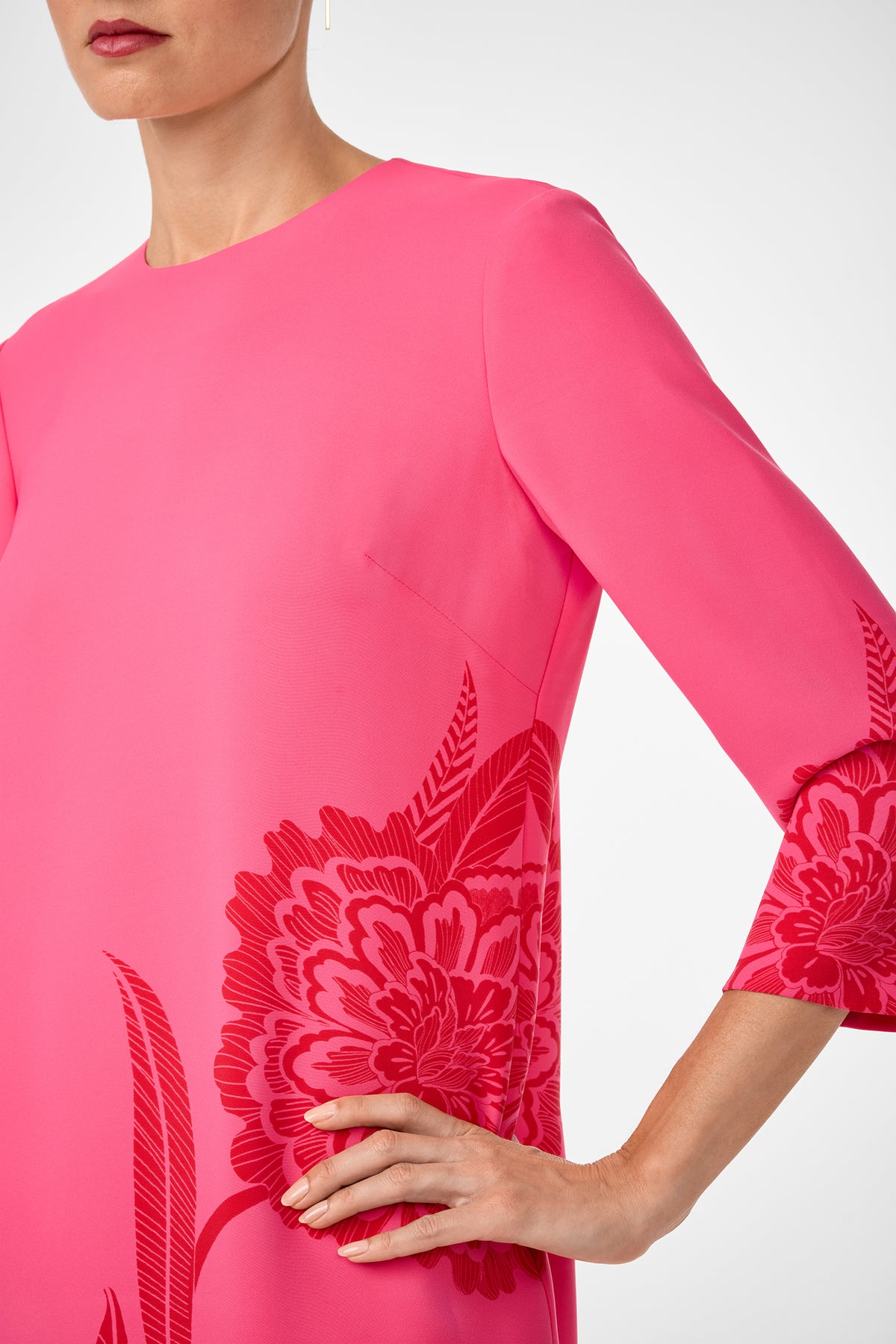 A woman in the Cointreau 2 Dress—a bright pink dress with red floral patterns and three-quarter sleeves—rests her hand on her hip, her face partially out of frame.