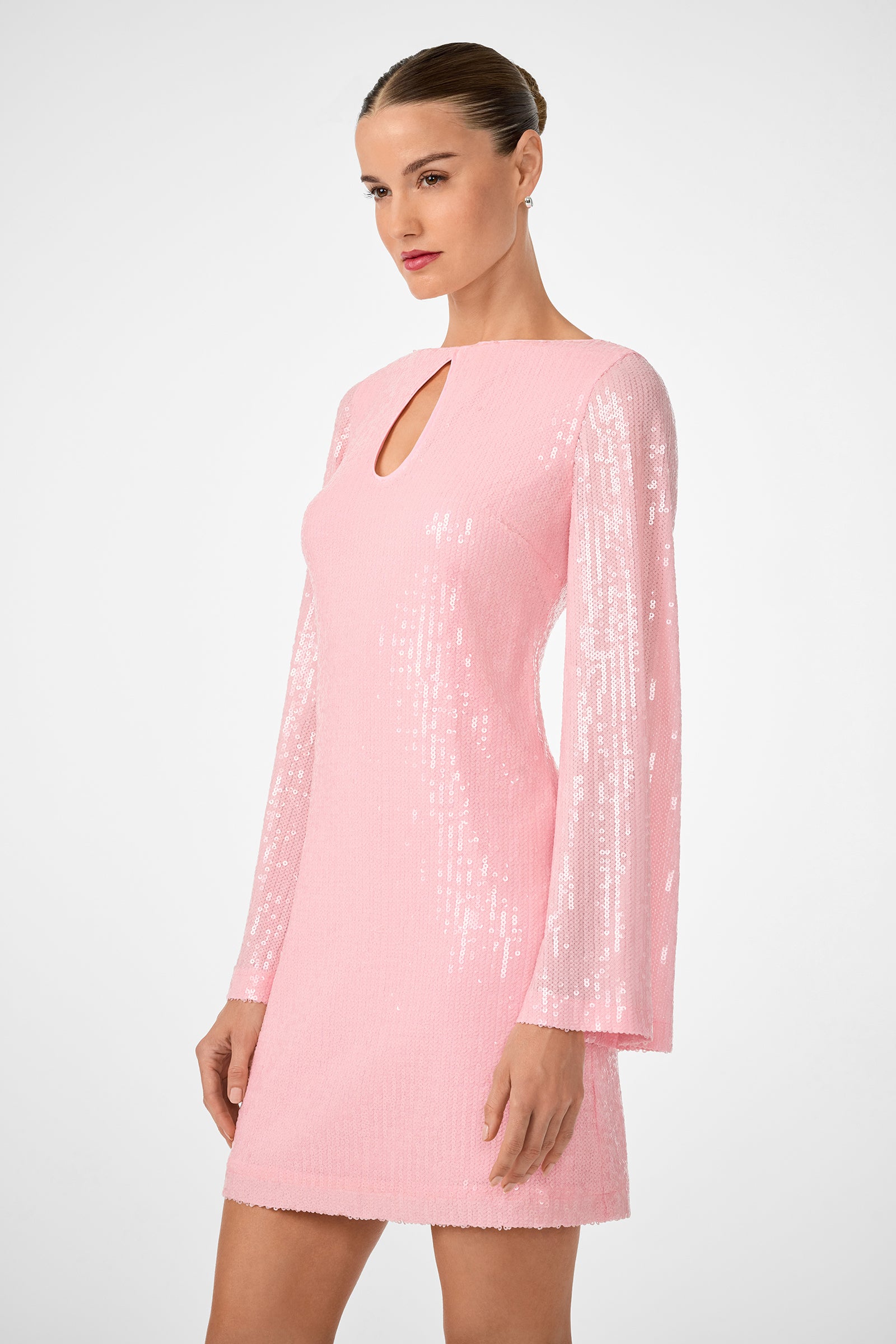 A woman stands against a plain light background, wearing the Singles 2 Dress—a light pink, long-sleeved sequined mini dress with a small keyhole neckline. Her hair is styled in a sleek bun.