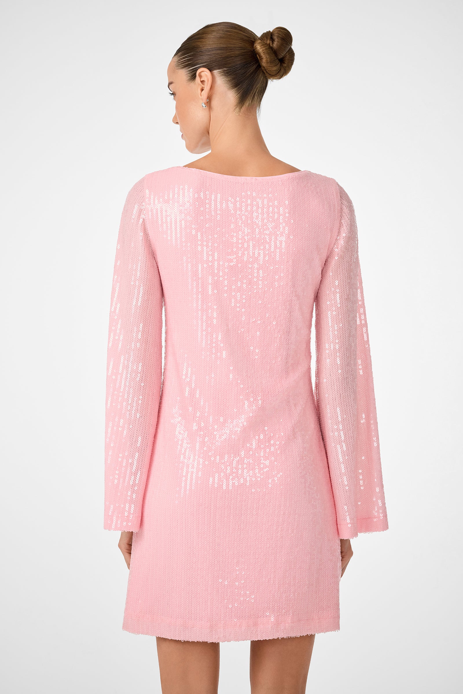 A woman with her hair in a neat bun is seen from behind wearing the Singles 2 Dress, a long-sleeved, light pink sequined mini dress. The background is plain white.