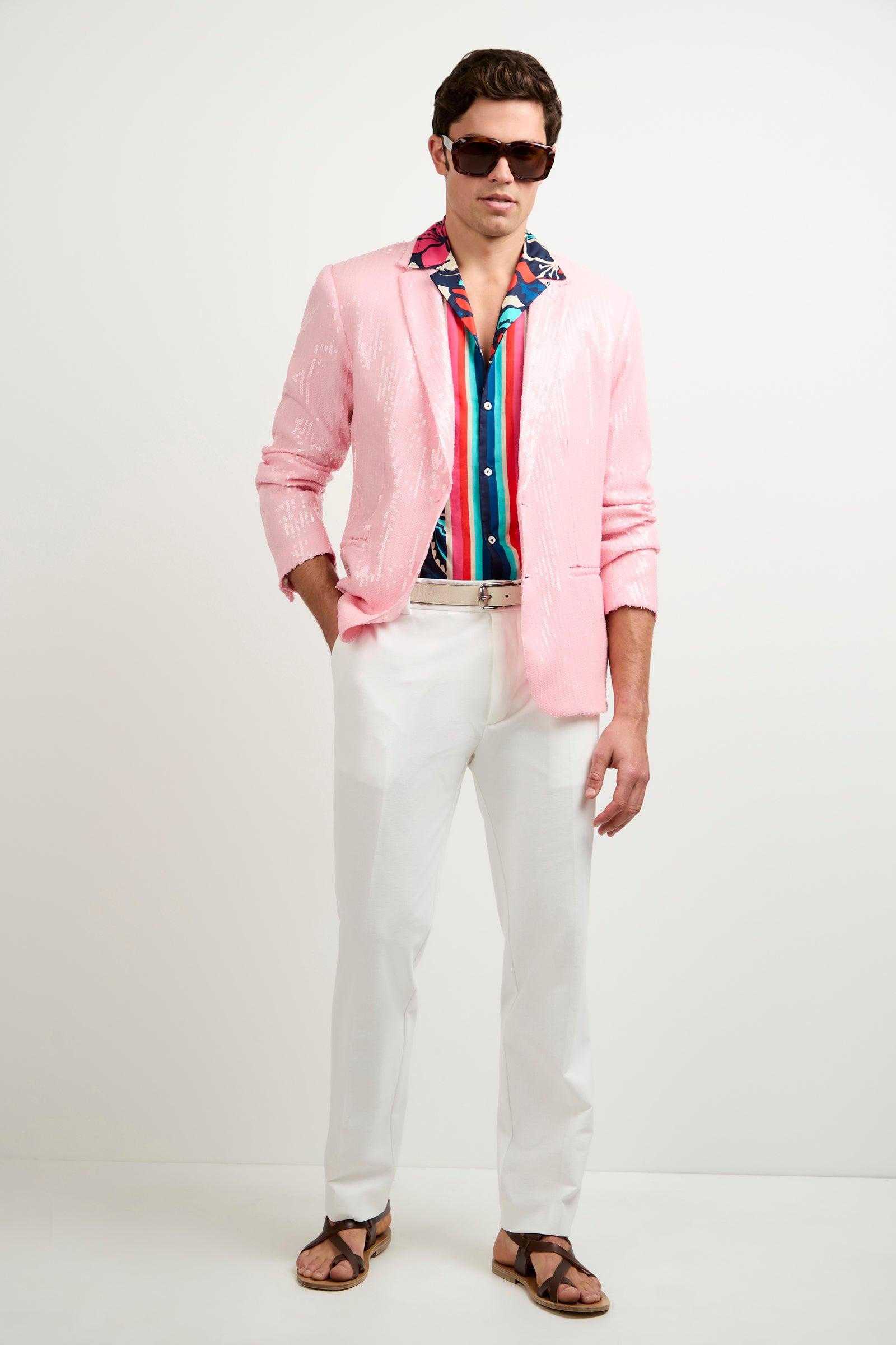 A man wearing the Bowie Sequin Blazer, a colorful striped shirt, white pants, dark sunglasses, and brown sandals stands against a plain white background.