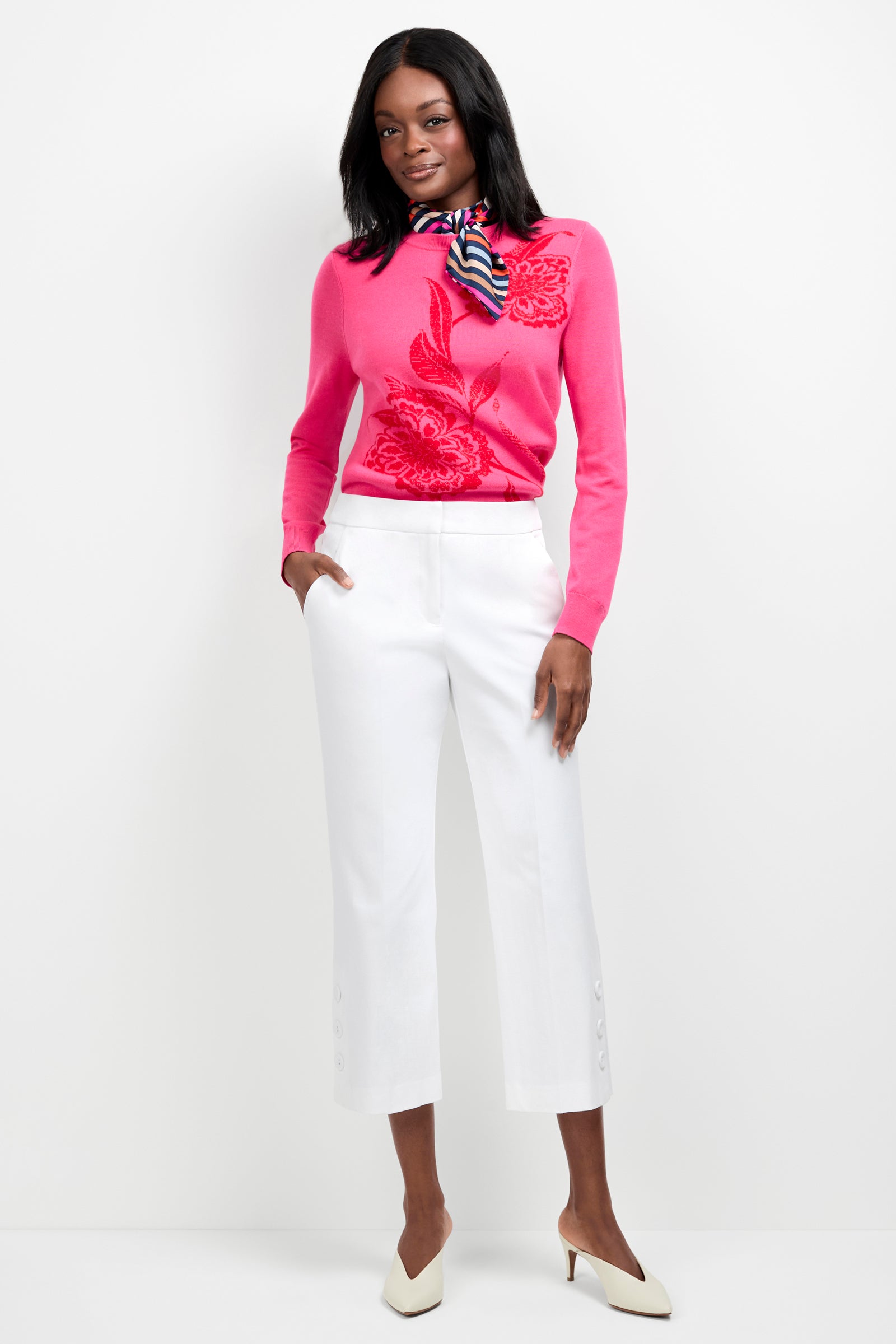A woman poses against a white background wearing the Old Fashioned Sweater in bright pink with a floral pattern, paired with white cropped pants with button hems, white heels, and a colorful striped scarf.