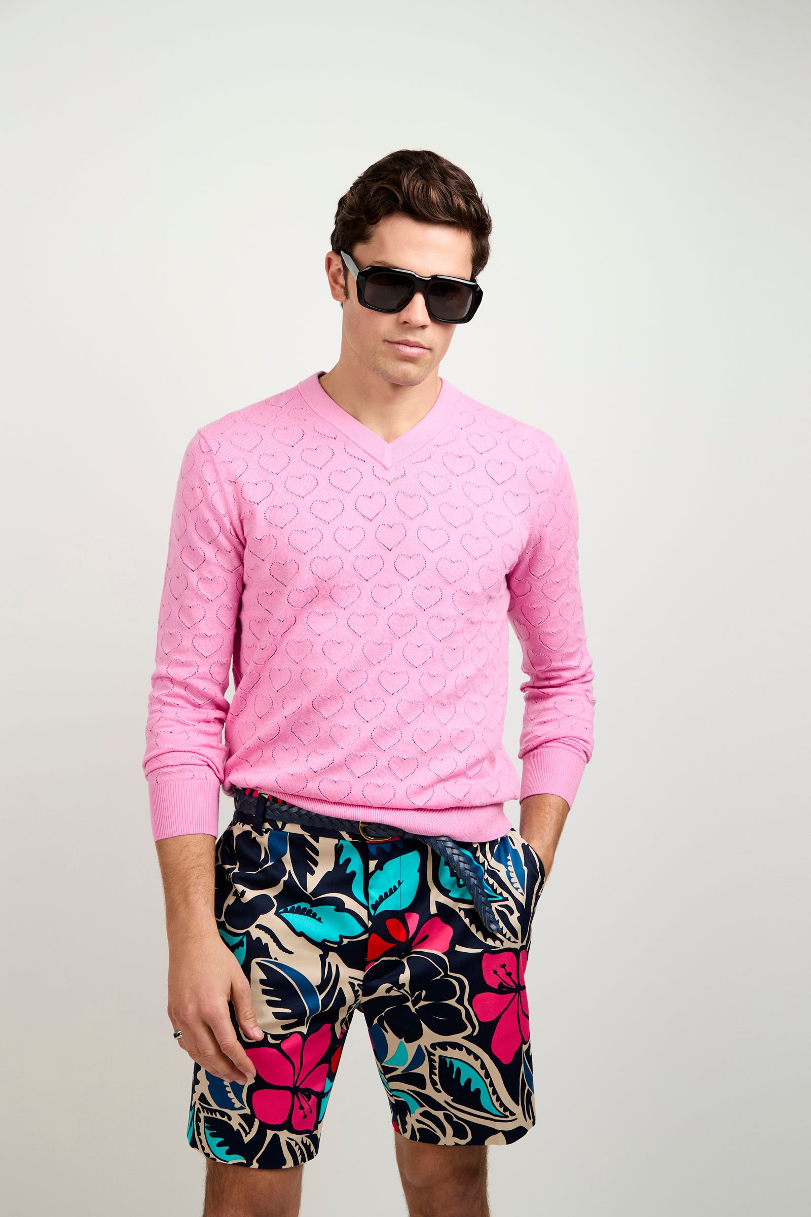 A man wears the Eros Heart Pointelle V-Neck Sweater, a light pink textured top, with black sunglasses and colorful floral shorts, standing against a plain light background with his hands in his pockets.