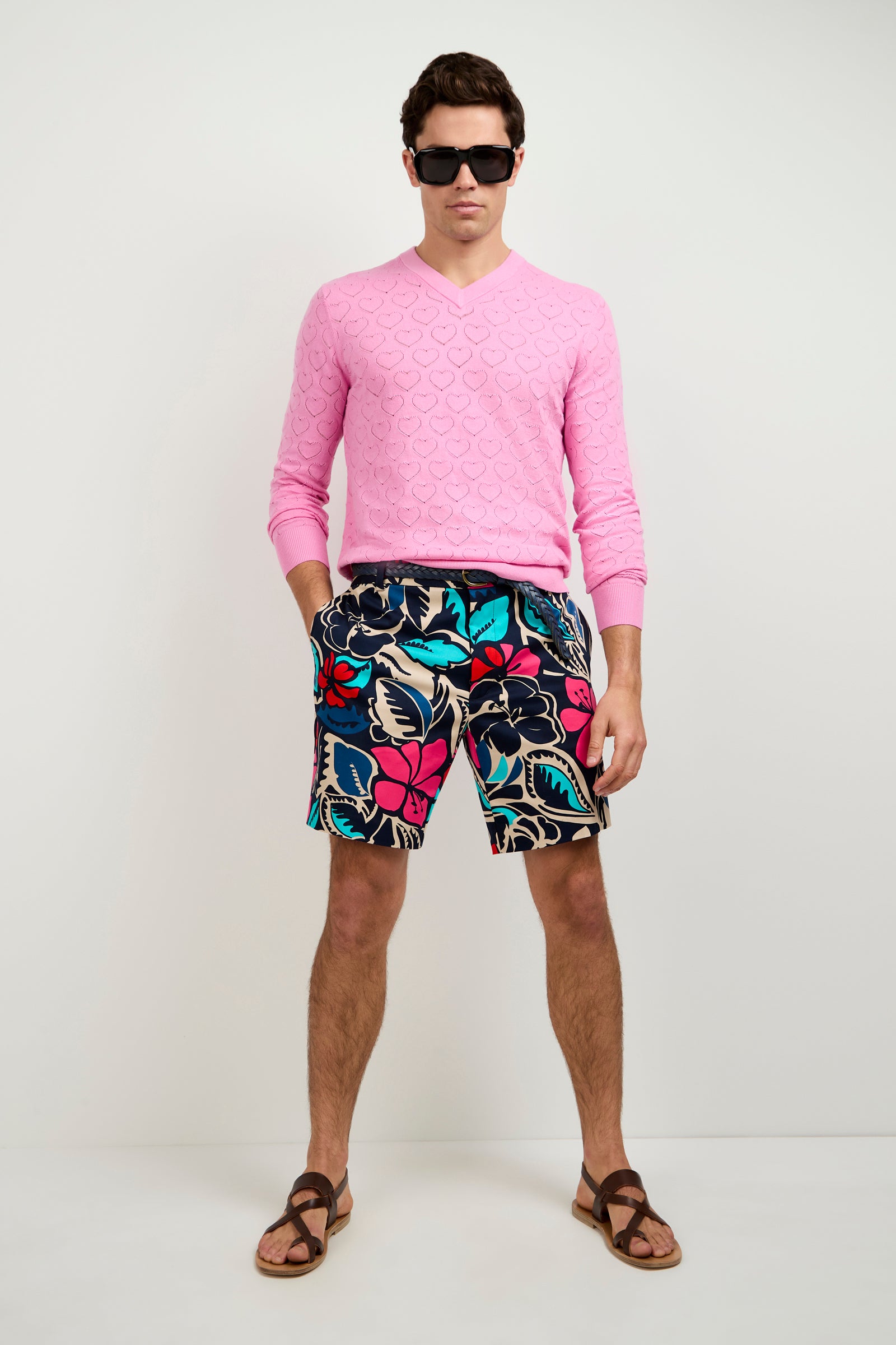 A man wearing dark sunglasses, the Eros Heart Pointelle V-Neck Sweater in light pink, colorful floral shorts, and brown sandals stands against a plain white background.