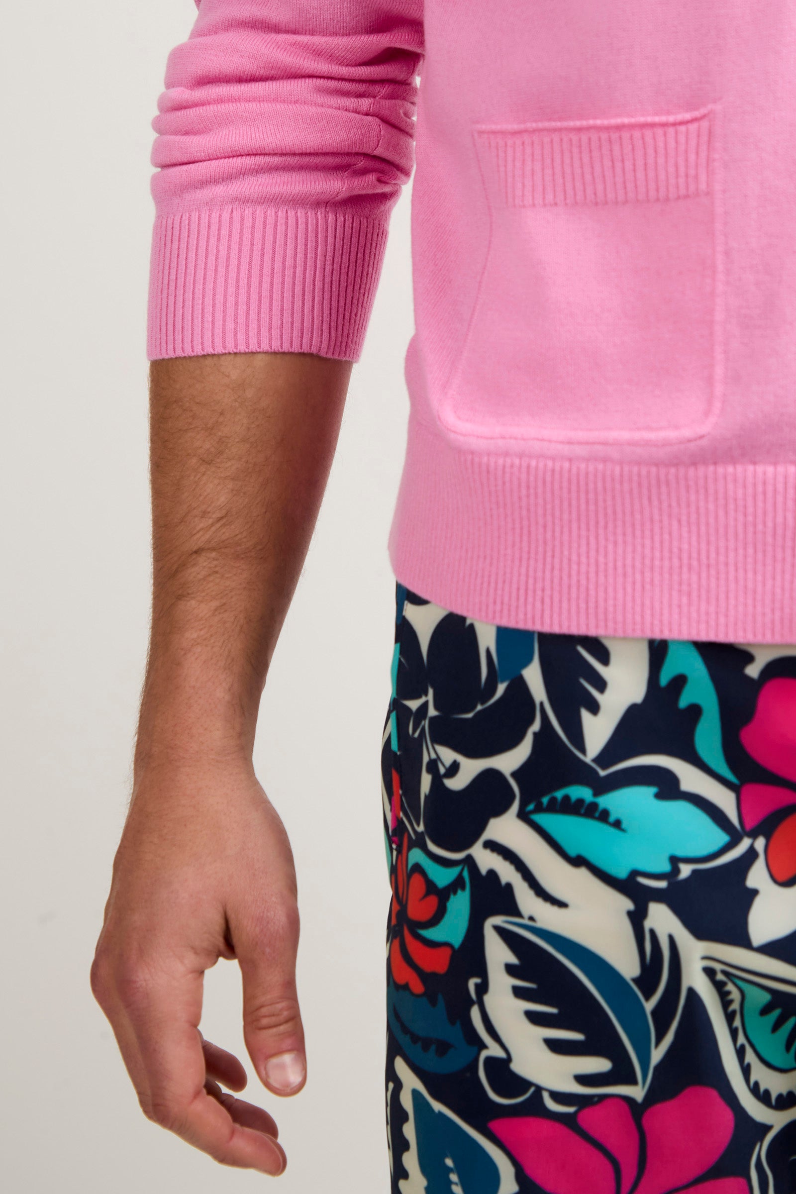 A person wearing a pink Jacques Cardigan with a pocket and colorful floral-patterned pants. Only the lower arm, hand, and part of the torso appear against a plain background.
