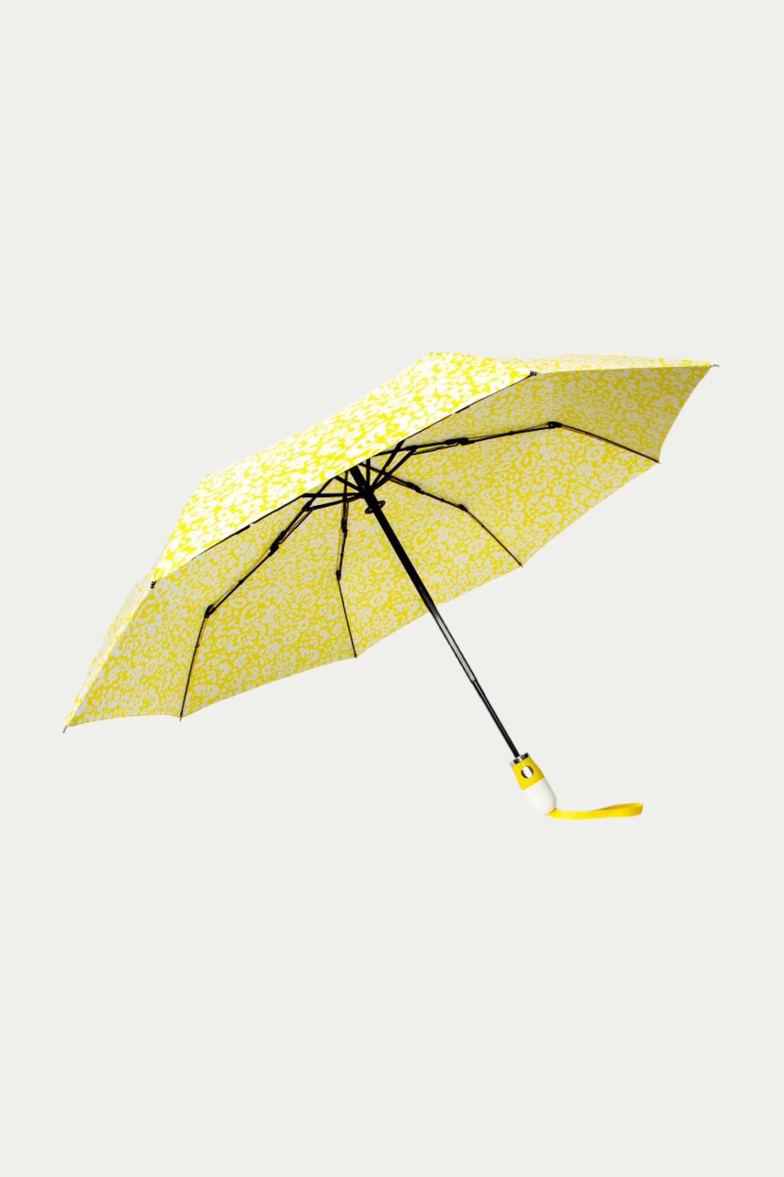 The TT X SR Compact Umbrella - Poolside Gossip in Desert Sun is open against a light background, showcasing its yellow and white floral pattern, black frame, yellow wrist strap, and white handle.