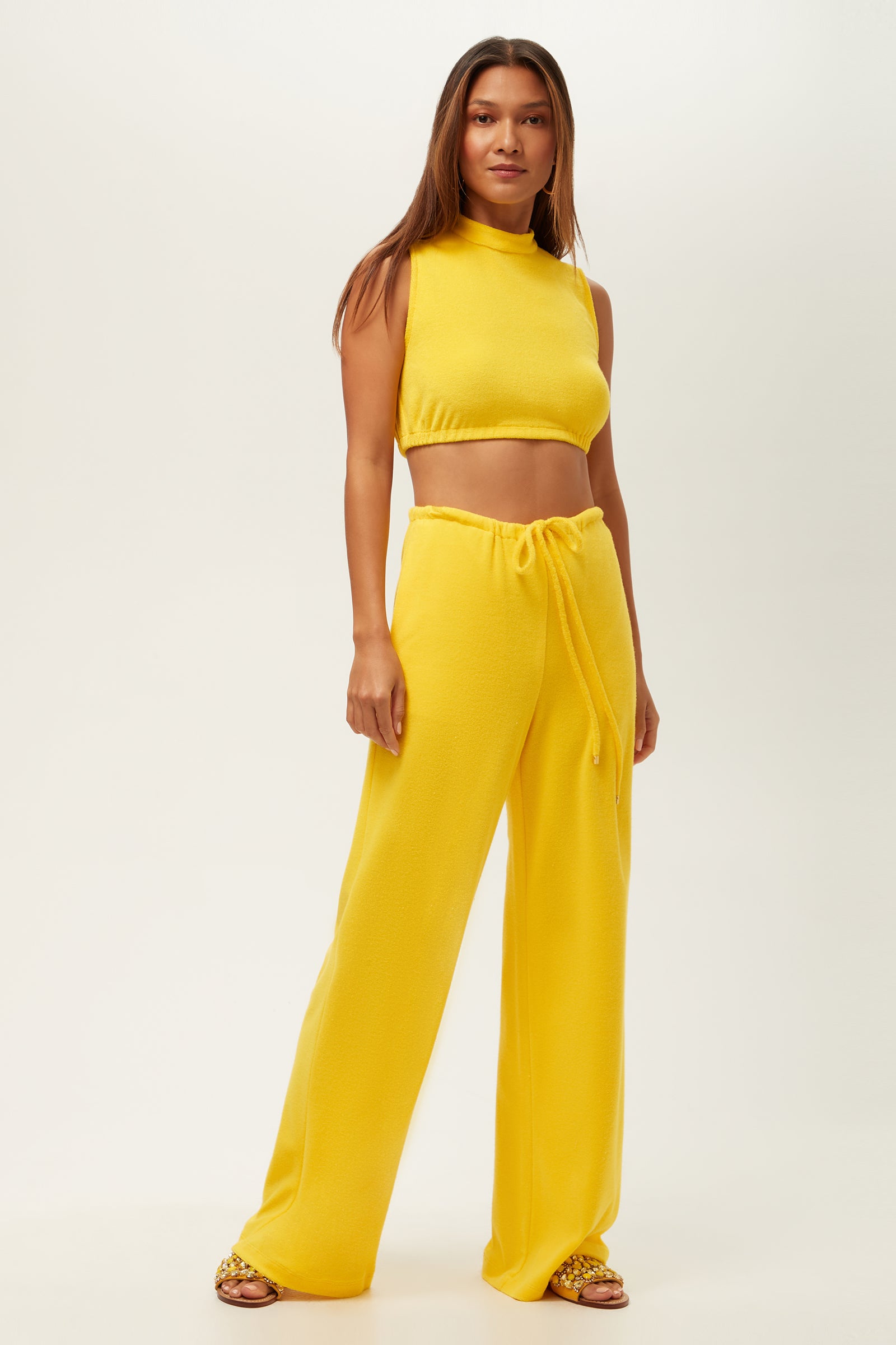 A woman with long brown hair stands against a plain background wearing the Linsk Wide-Leg Pant - Poolside Terry, paired with a sleeveless yellow crop top and slide sandals.