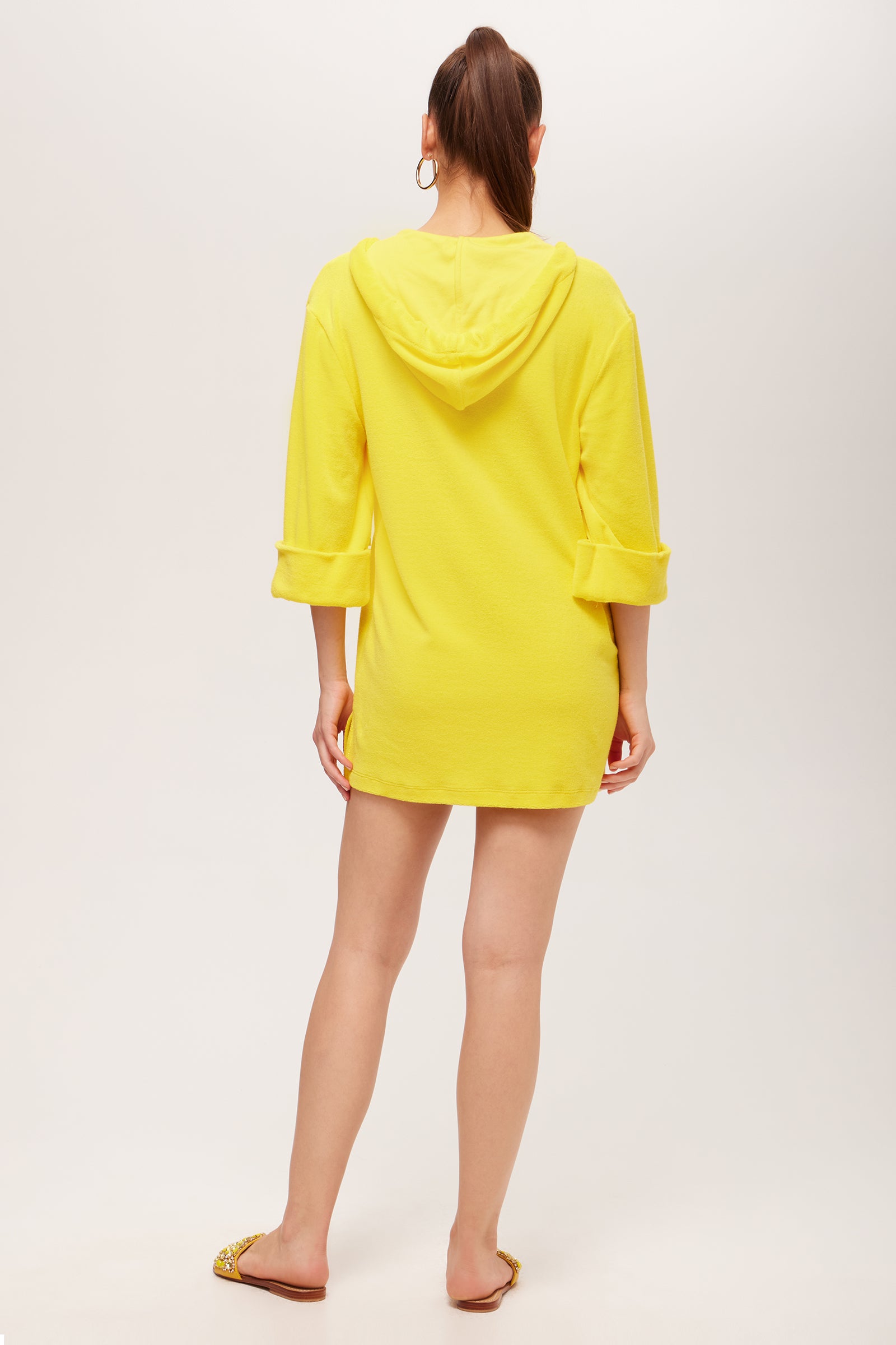 A woman with a high ponytail faces away, wearing the Poolside 3/4-Sleeve Hoodie in bright yellow terry as a dress with rolled-up sleeves and yellow sandals, set against a plain light background.