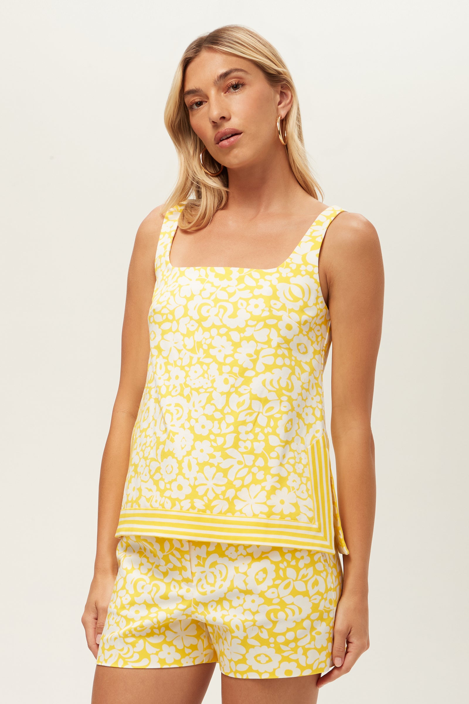 A woman with long blonde hair wears the Aspire 2 Sleeveless Top - Poolside Gossip in Desert Sun, featuring a yellow and white floral pattern and square neckline. She stands against a plain background, looking relaxed with one arm by her side.
