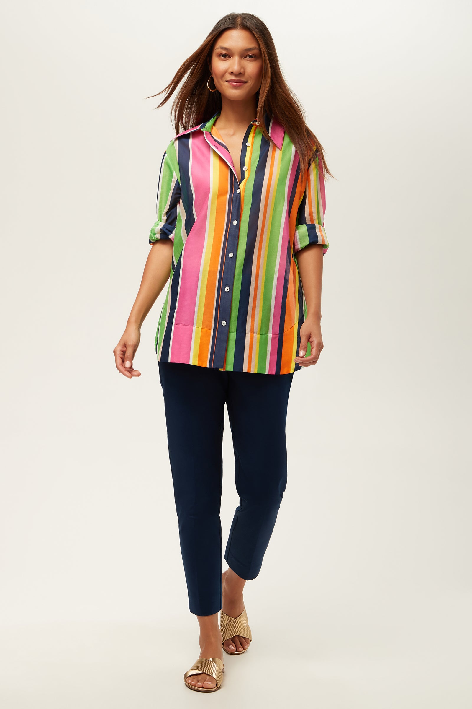 A woman walks confidently wearing the Swing Short-Sleeve Top in Splash Stripe with navy blue pants and gold sandals, set against a plain white background.
