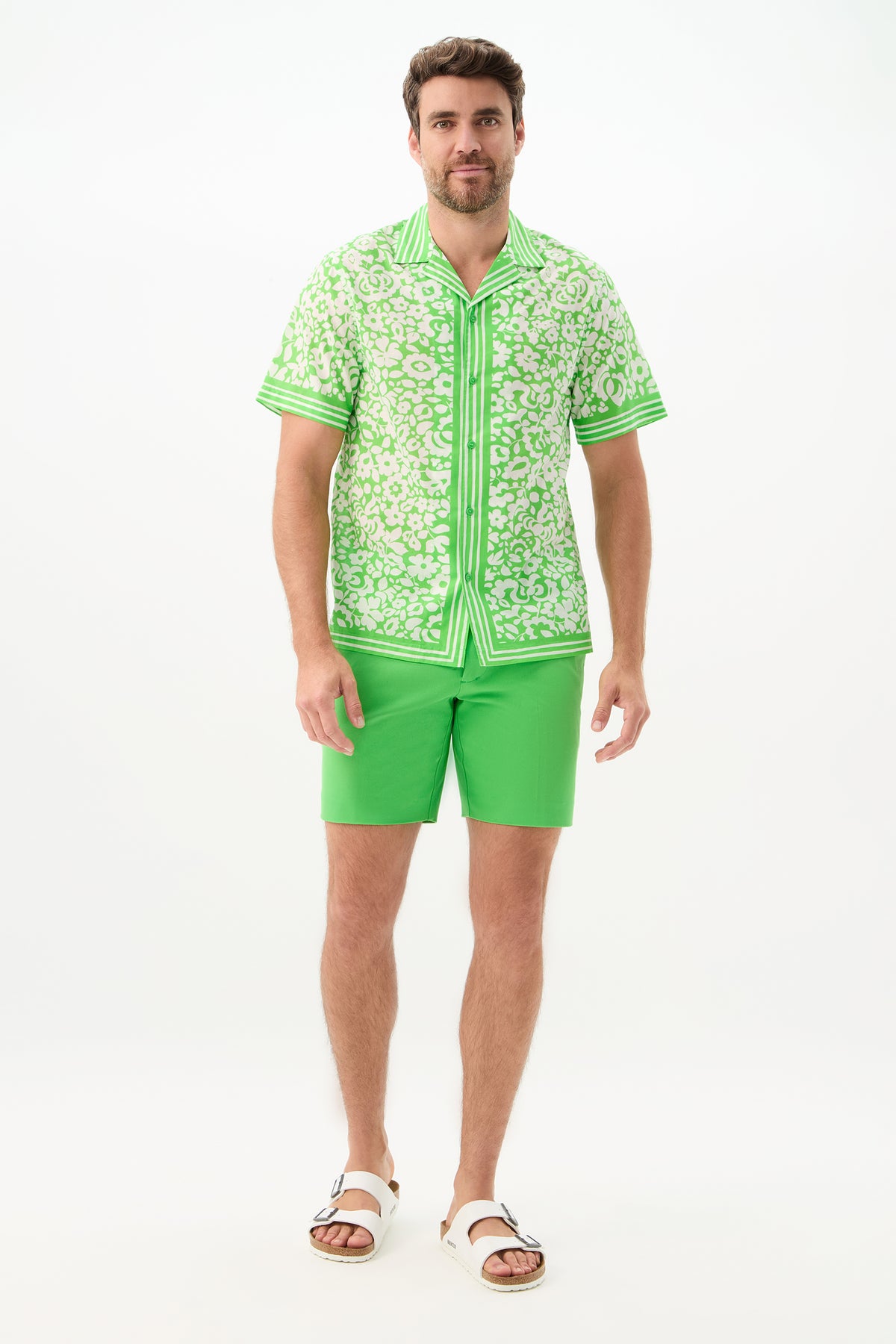 A man with short brown hair and a beard wears the Cyrus Short - Cotton Bi-Stretch in solid green, a green patterned short-sleeve shirt, and white sandals, standing against a plain white background.