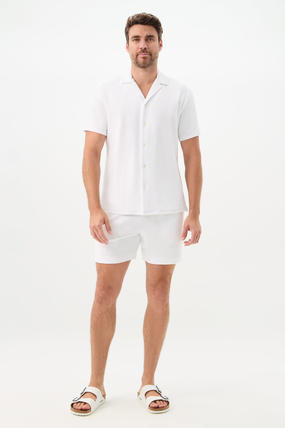 A man with short brown hair and a trimmed beard stands against a plain white background, wearing the Felix Short-Sleeve Knit Shirt - Linsk Terry, paired with matching white shorts and white sandals.