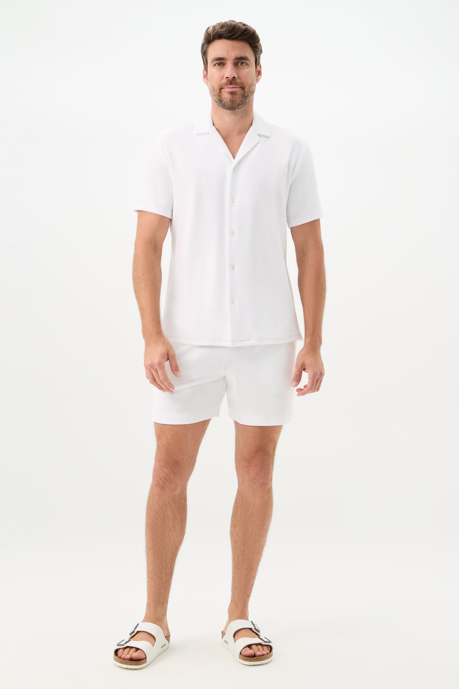 A man with short brown hair and a trimmed beard stands against a plain white background, wearing the Felix Short-Sleeve Knit Shirt - Linsk Terry, paired with matching white shorts and white sandals.