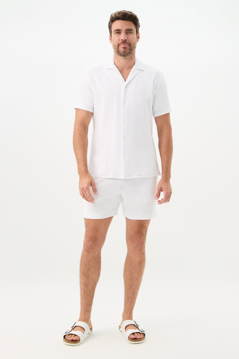 A man stands against a plain white background wearing a white short-sleeve button-up shirt, Lounge Knit Short - Linsk Terry in white, and white sandals.
