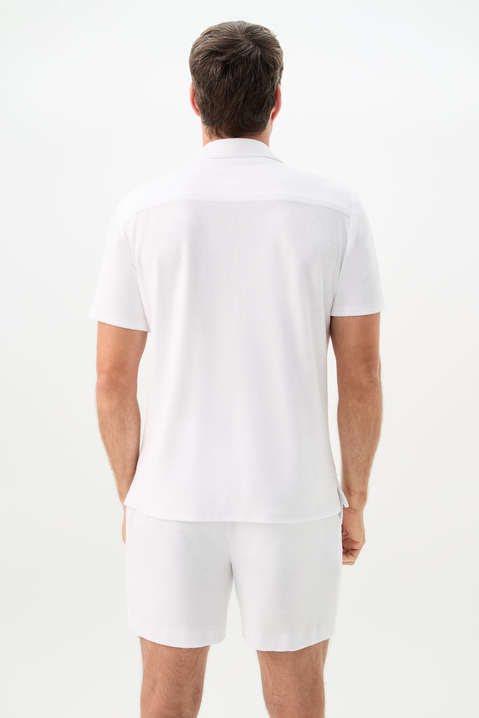 A man with short brown hair stands with his back to the camera, wearing the Felix Short-Sleeve Knit Shirt - Linsk Terry and white shorts against a plain white background.