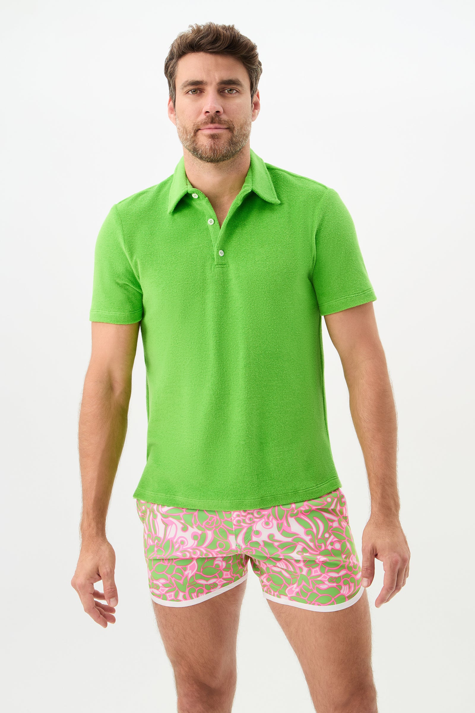 A man with short brown hair and a beard wears the Dallas Short-Sleeve Polo Shirt - Linsk Terry in Serene Green, paired with pink patterned shorts, standing against a plain white background.