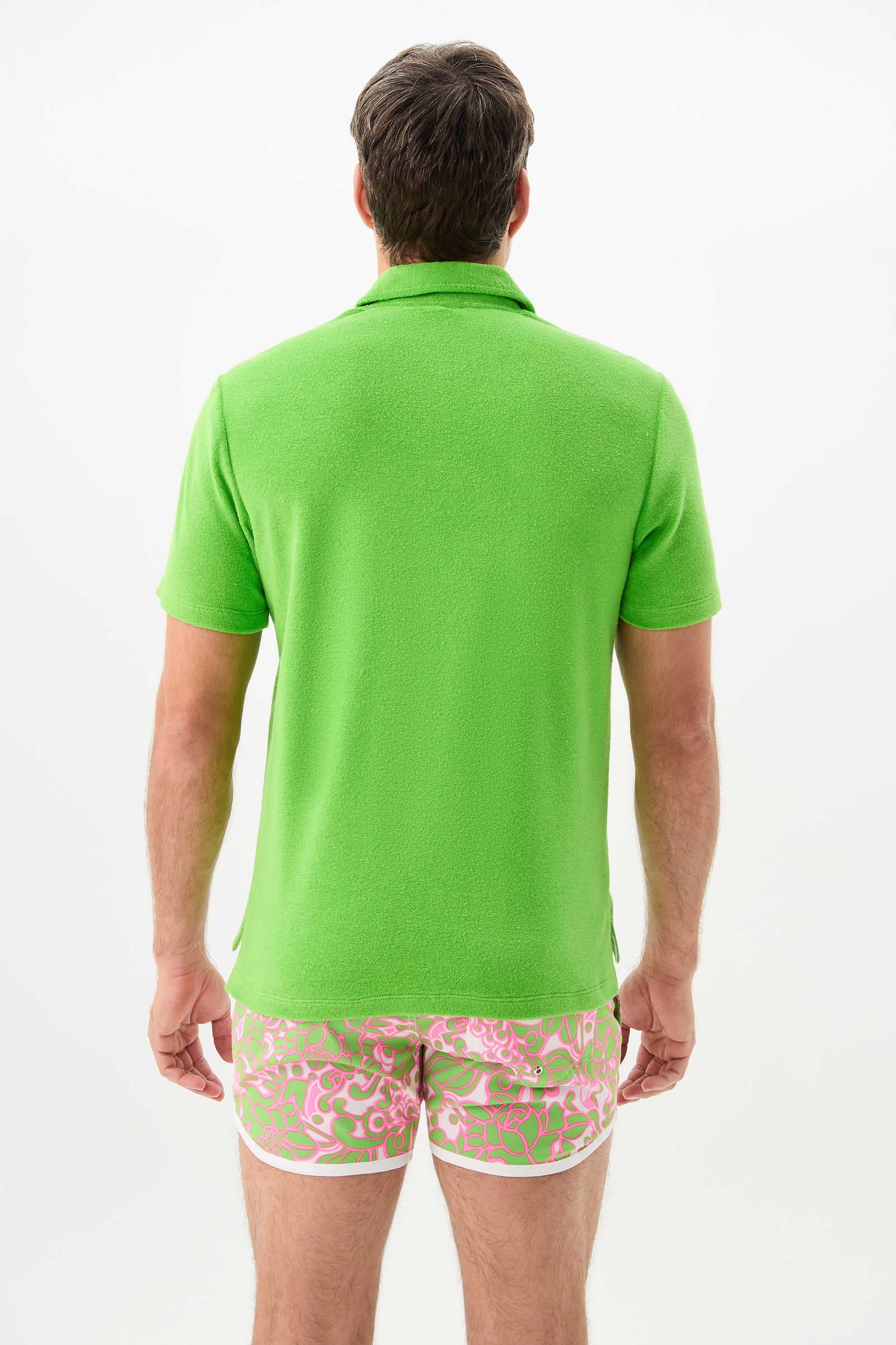 A person stands with their back to the camera, wearing the Dallas Short-Sleeve Polo Shirt - Linsk Terry in bright green and pink shorts with a green and white abstract pattern, set against a plain white background.