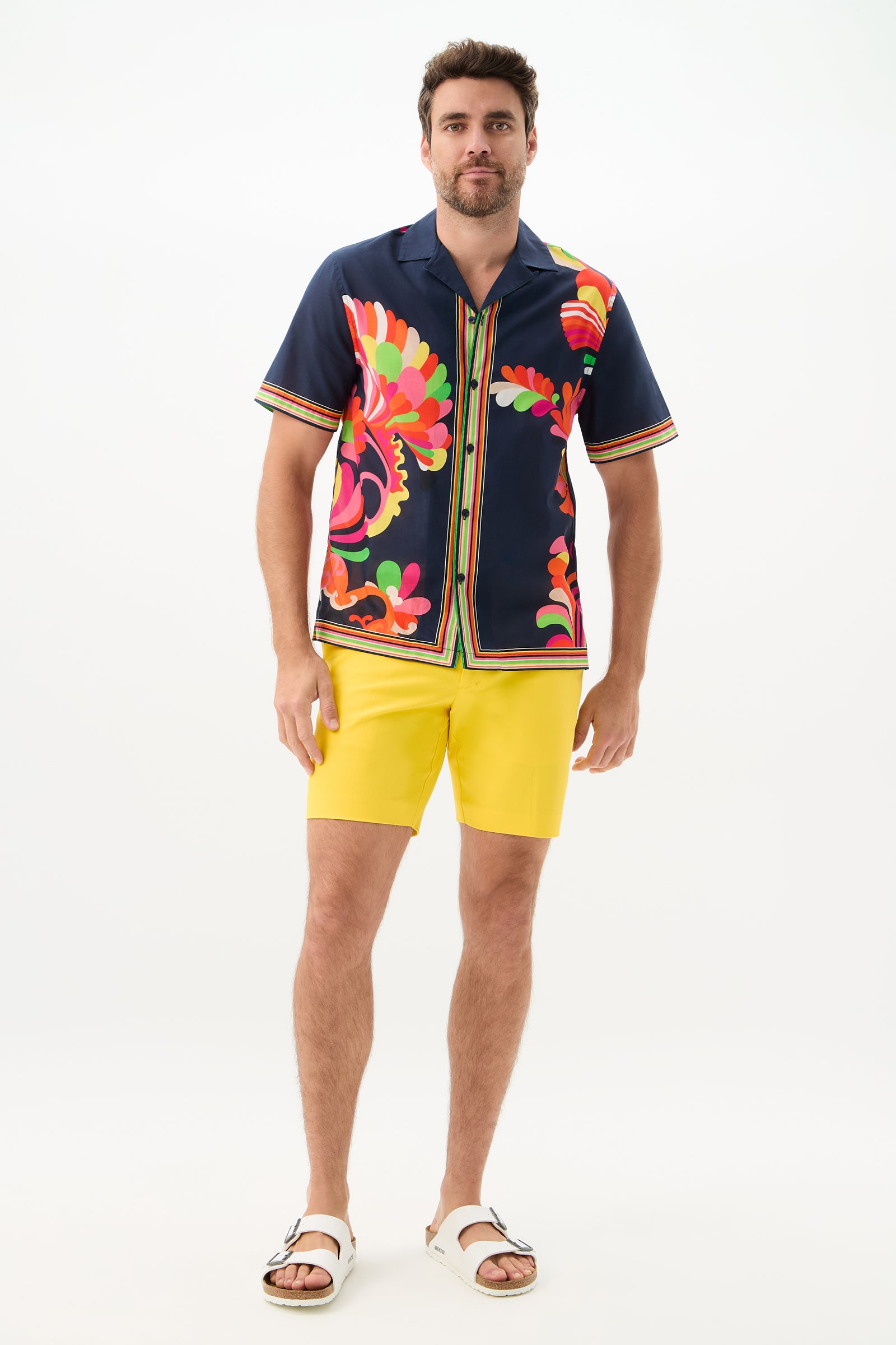 A man with short dark hair and a beard stands facing forward against a plain white background, wearing the Josue Short-Sleeve Shirt - Palms Paradise, yellow shorts, and white sandals.
