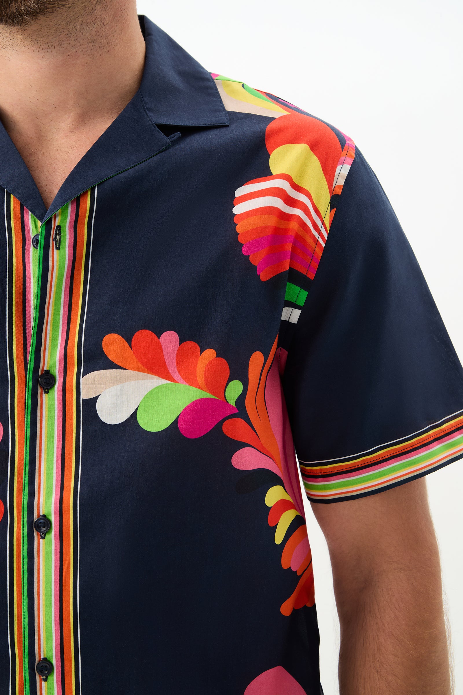 A person wearing the Josue Short-Sleeve Shirt - Palms Paradise, featuring navy blue fabric with colorful abstract patterns and vertical multicolored stripes; image cropped to show the upper torso and part of the left arm.