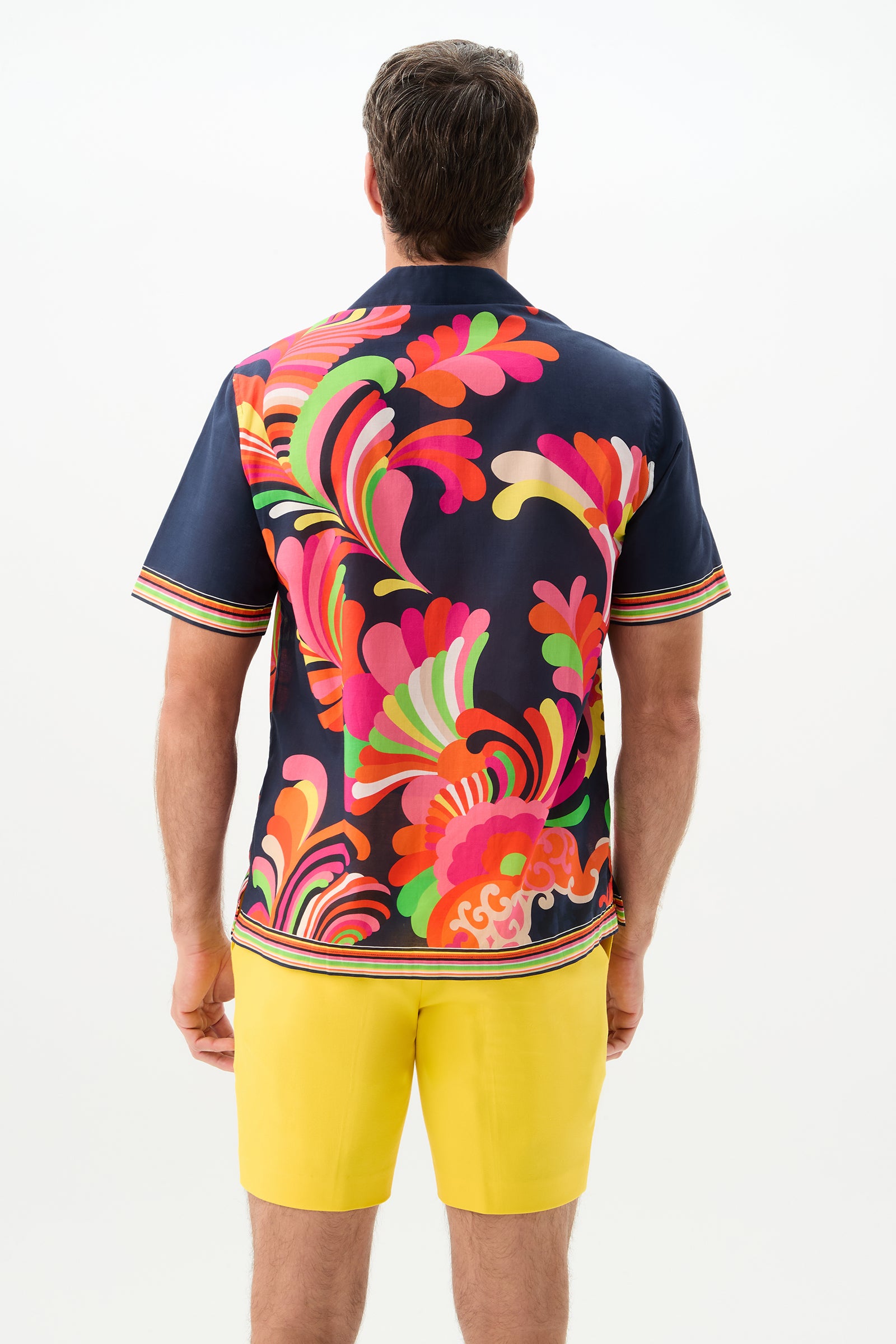 A man faces away, wearing the Josue Short-Sleeve Shirt - Palms Paradise with bold, colorful swirl patterns and bright yellow shorts against a plain white background.