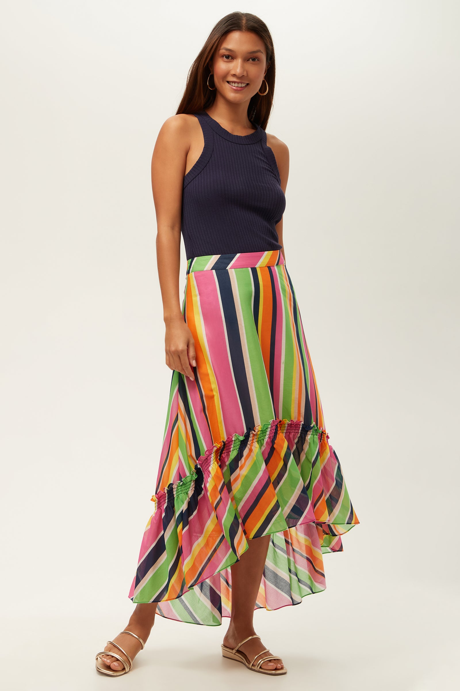 A woman smiles against a plain light background, wearing the Rosamund 2 High-Low Skirt in Splash Stripe with a ruffled hem, a sleeveless navy ribbed top, and gold sandals.