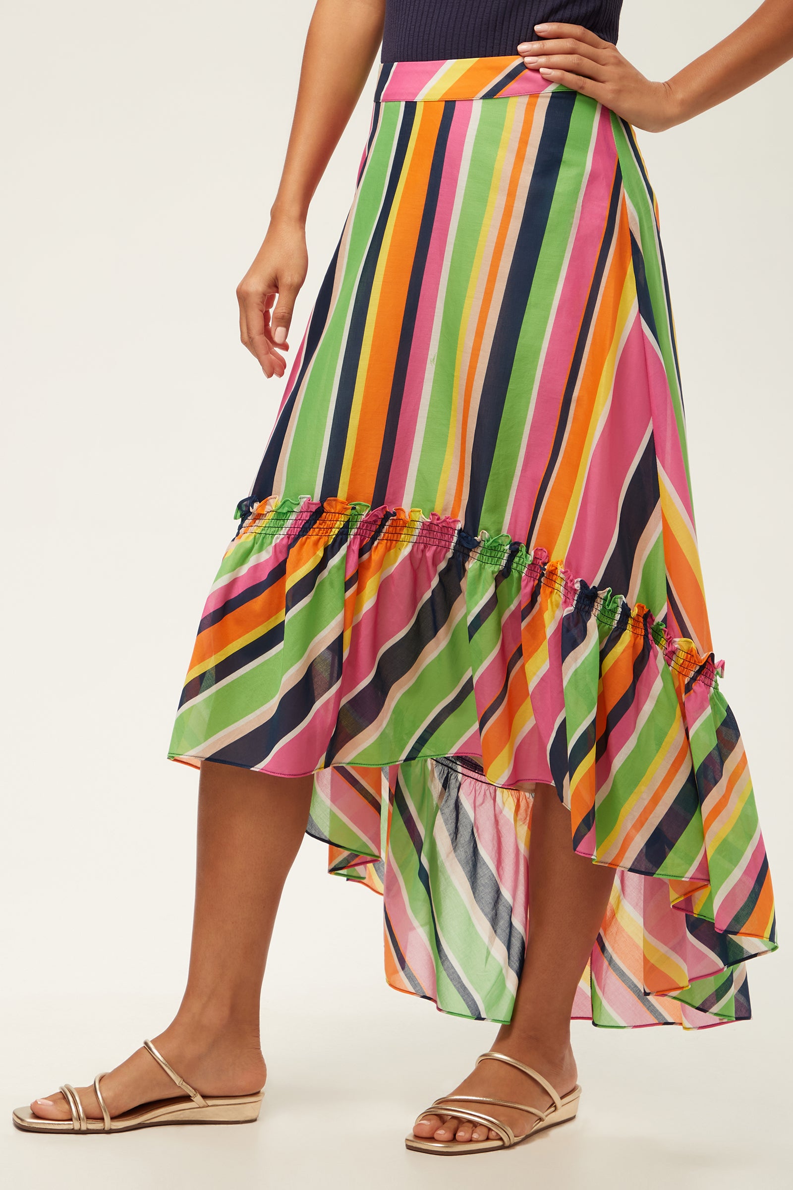 A person wearing the Rosamund 2 High-Low Skirt - Splash Stripe, featuring colorful diagonal stripes in green, pink, orange, blue, and yellow with a ruffled hem, paired with flat nude sandals.