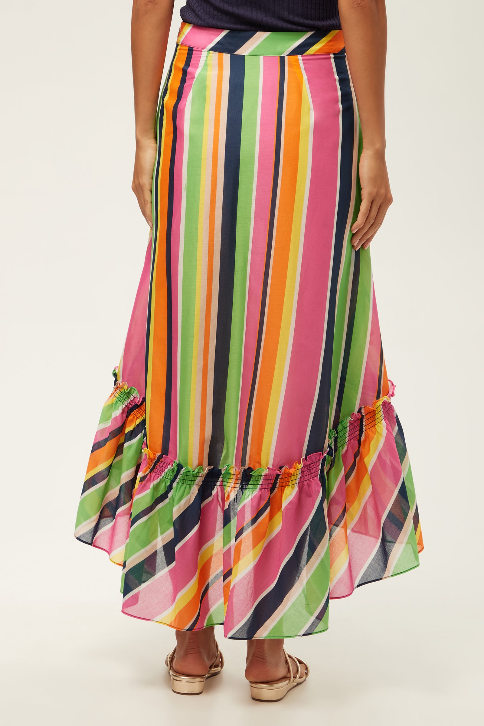 A person faces away, wearing the Rosamund 2 High-Low Skirt - Splash Stripe, a colorful, high-waisted maxi with vertical pink, orange, green, yellow, and blue stripes and a ruffled hem. They pair it with sandals.