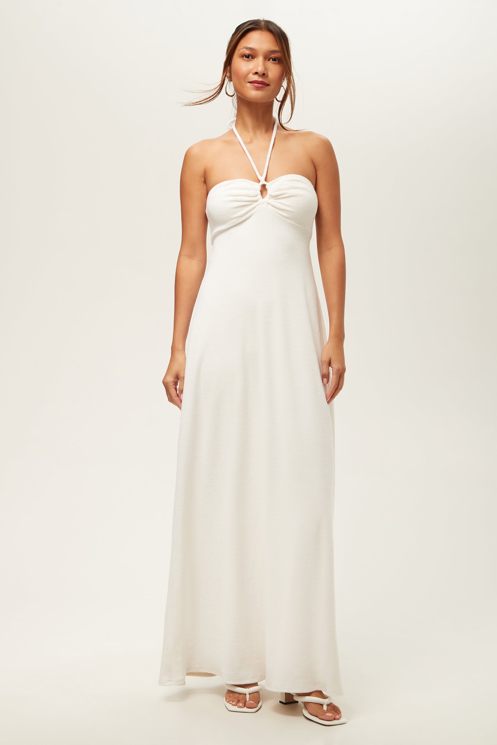 A woman wears the Palm Sleeveless Maxi Dress - Poolside Terry in white, featuring a halter neck and chest keyhole cutout, paired with white open-toe sandals. She stands against a plain, light-colored background.