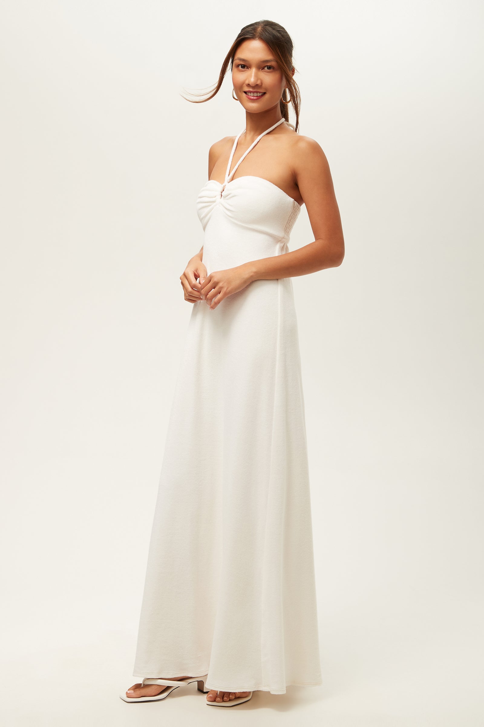 A woman smiles in the Palm Sleeveless Maxi Dress - Poolside Terry, featuring a long, white halter-neck style with gathered bust detail. She wears white sandals and has her hair in a loose updo, set against a plain white background.