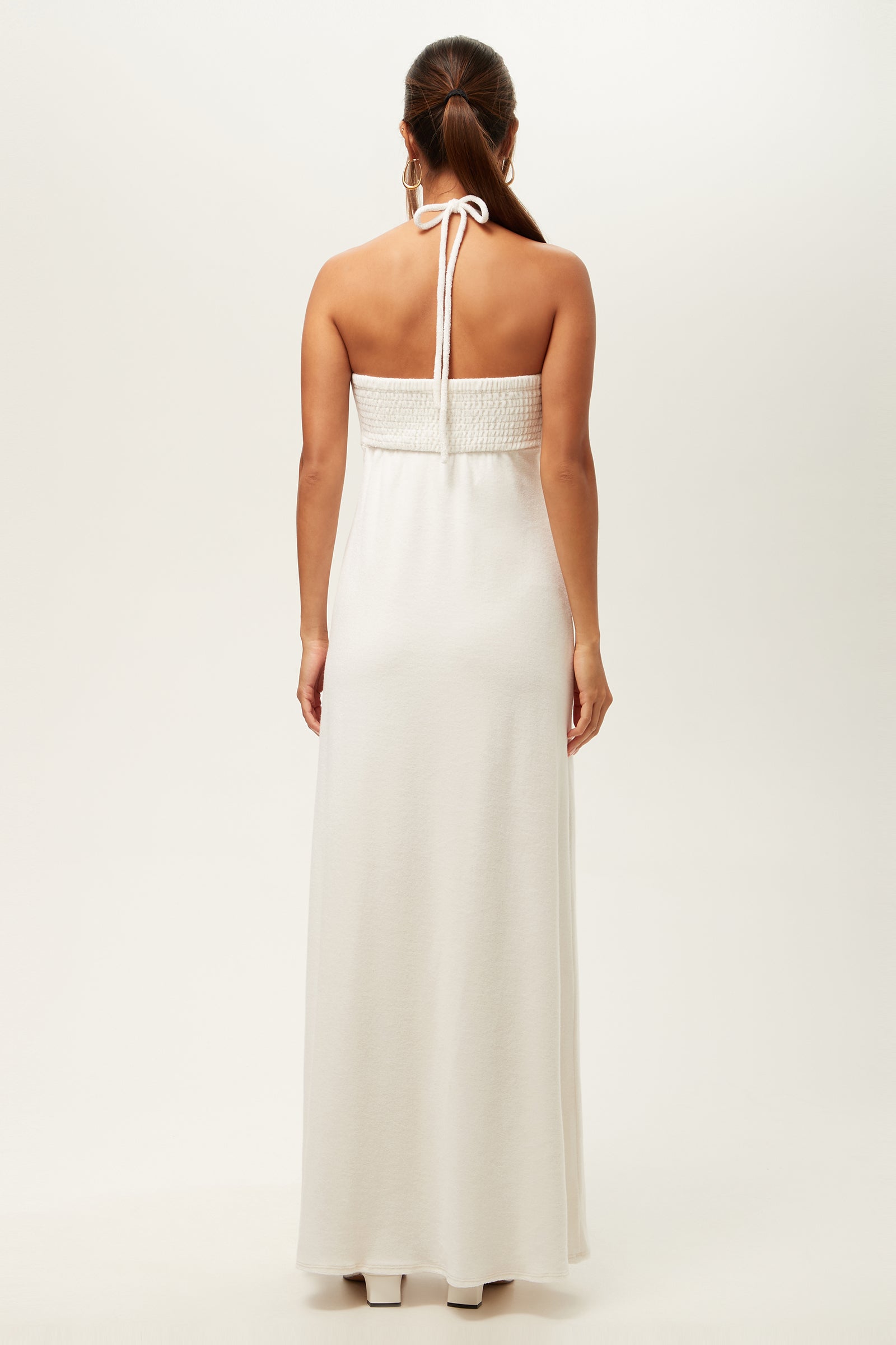 A woman with brown hair in a ponytail wears the Palm Sleeveless Maxi Dress - Poolside Terry, a long white halter-neck style. Seen from behind, she shows the neck tie and smocked back against a plain, light background.