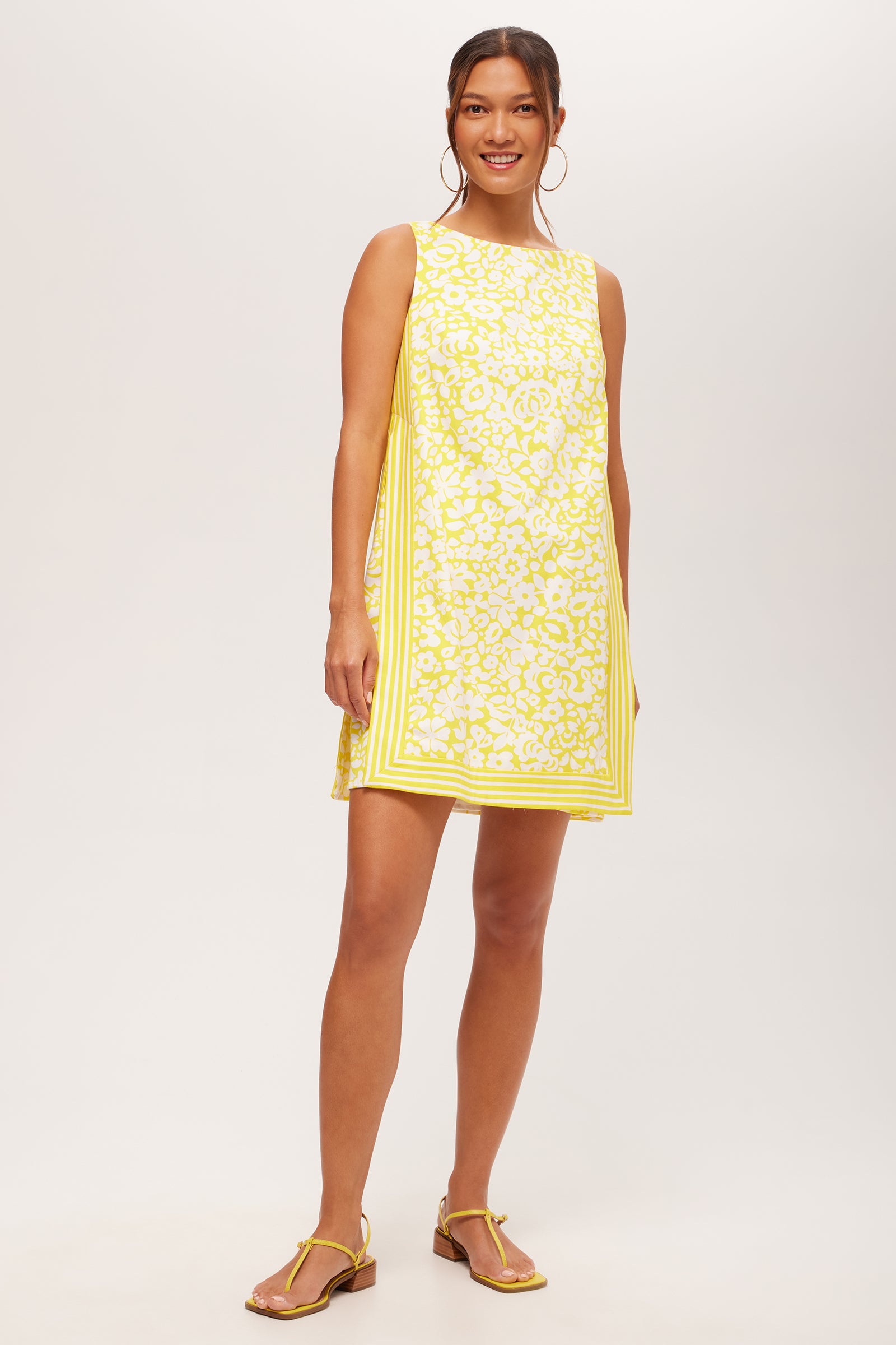 A woman smiles in the Brynne 3 Sleeveless Dress - Poolside Gossip in Desert Sun, featuring a square neckline and yellow trim. She pairs it with yellow sandals and hoop earrings against a plain background.