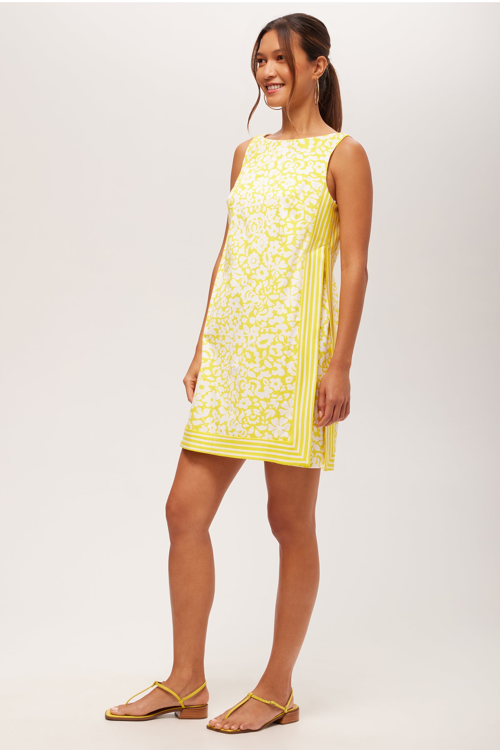 A woman smiles, wearing the Brynne 3 Sleeveless Dress - Poolside Gossip, a yellow and white patterned shift dress with side pleats and striped borders. She has long hair tied back and pairs the look with flat yellow sandals.
