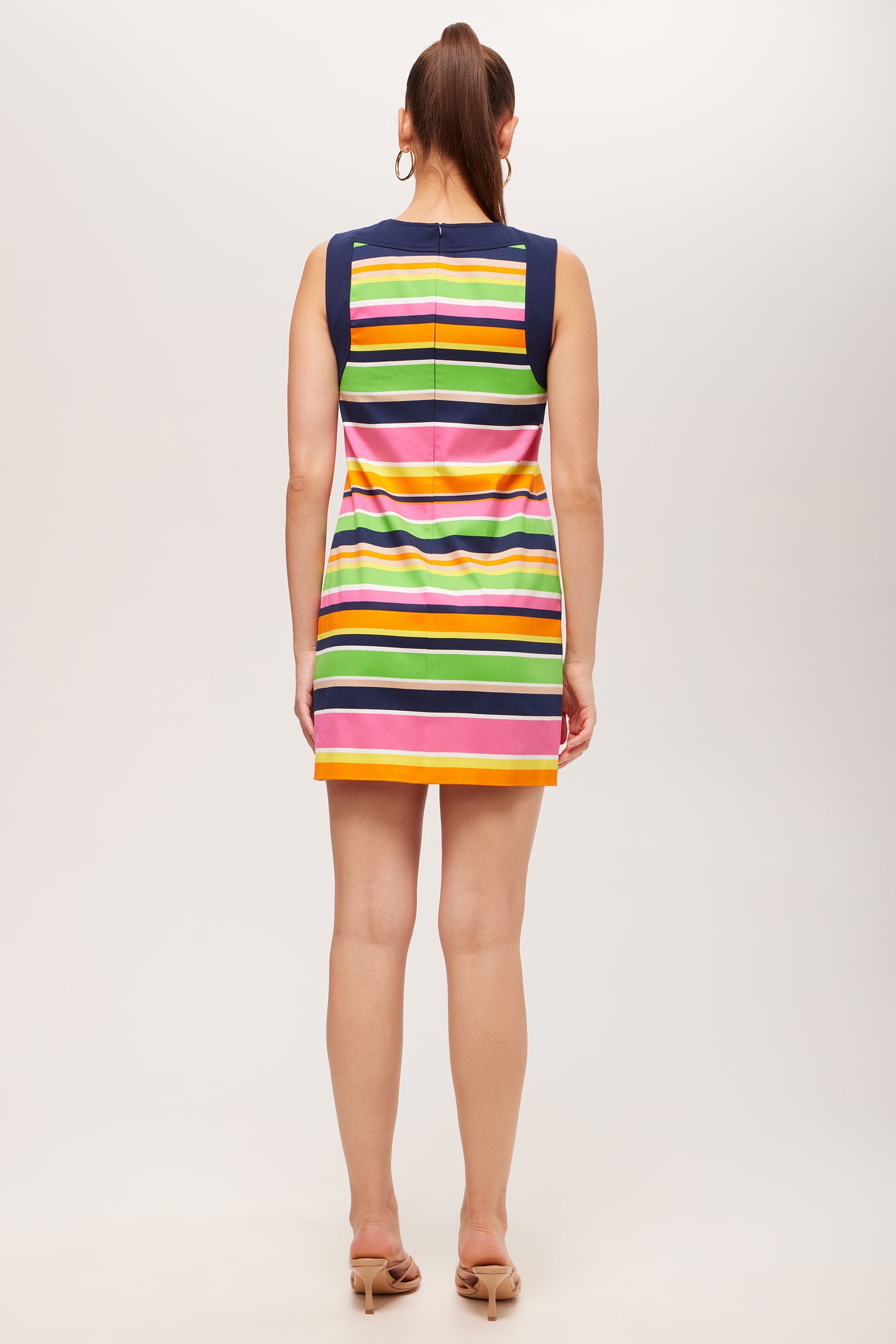 A woman with medium-length brown hair in a ponytail wears the Bev Sleeveless Cutout Dress - Splash Stripe, featuring multicolored horizontal stripes in yellow, green, navy, pink, and white. She pairs it with nude heeled sandals.