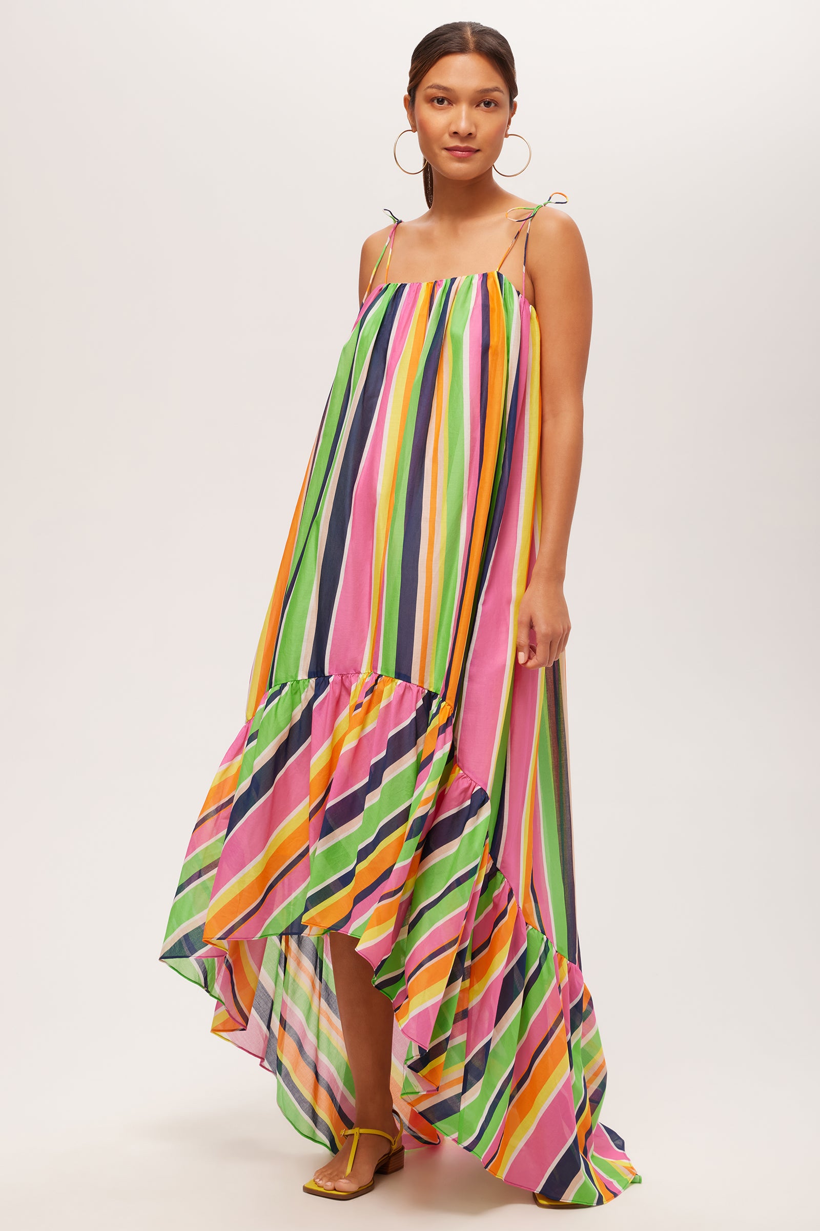 A woman wears the Enchant 2 Sleeveless Dress - Splash Stripe in Multi, featuring thin straps, a colorful striped pattern, and a ruffled high-low hem. She pairs it with hoop earrings and sandals for a vibrant look.