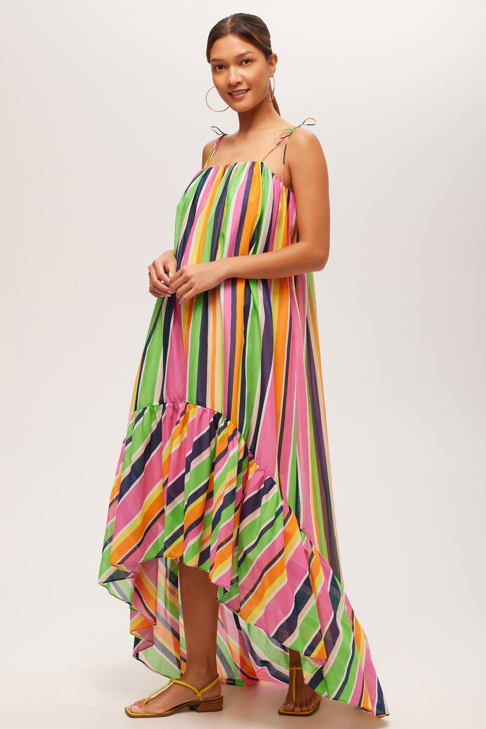 A woman smiles in the Enchant 2 Sleeveless Dress – Splash Stripe, a loose, high-low maxi featuring vibrant pink, green, orange, yellow, and blue stripes. She accessorizes with gold hoop earrings and flat sandals.