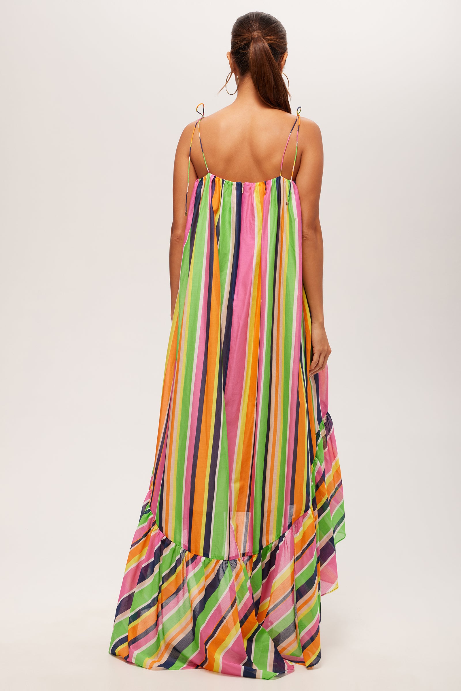A woman with long brown hair, seen from the back, wears the Enchant 2 Sleeveless Dress - Splash Stripe, a colorful maxi dress with thin shoulder straps, vertical green, yellow, pink, purple, and orange stripes, and a ruffled hem.