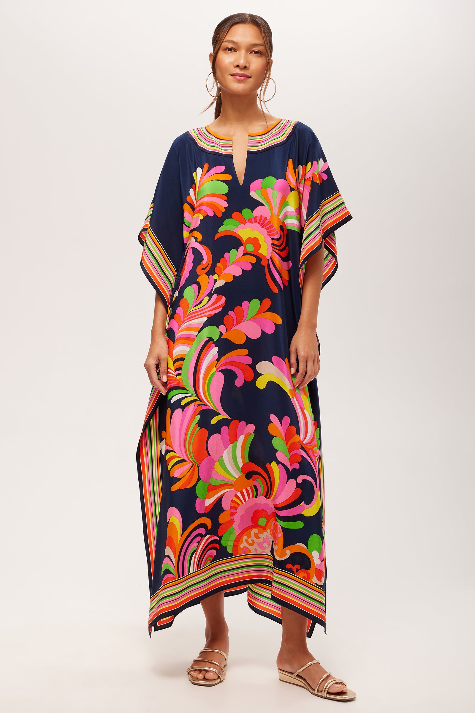 A woman stands facing forward, wearing the Theodora Maxi Caftan - Paradise Palms Silk in Multi. Her hair is tied back, and she accessorizes with hoop earrings and sandals.