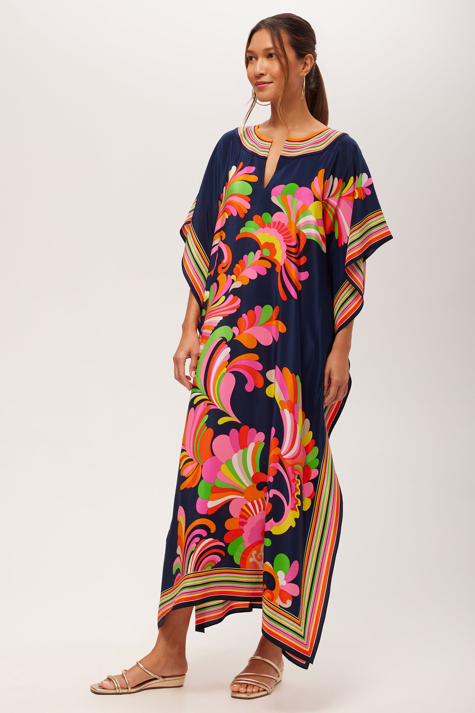 A woman wears the Theodora Maxi Caftan - Paradise Palms Silk, a loose navy blue caftan featuring vibrant floral and swirl patterns in pink, orange, green, and yellow. She pairs it with flat sandals against a plain white background.